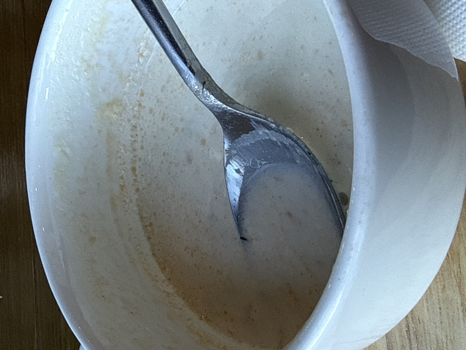 This image depicts a close-up, top-down view of a partially consumed creamy liquid within a white ceramic bowl. A silver-colored metal spoon rests inside the bowl, its bowl submerged in the liquid, with the handle extending towards the right. Residue and small flecks, possibly of a darker substance, adhere to the inner walls of the bowl above the liquid line. A small, dark, elongated object, consistent with an insect such as a fly, is visible on the surface of the liquid near the tip of the spoon. The bowl is positioned on a light brown, possibly wooden surface, with a white, textured material resembling a paper napkin visible to the right of the bowl. This scene is located in Shlisselburg, Russia.