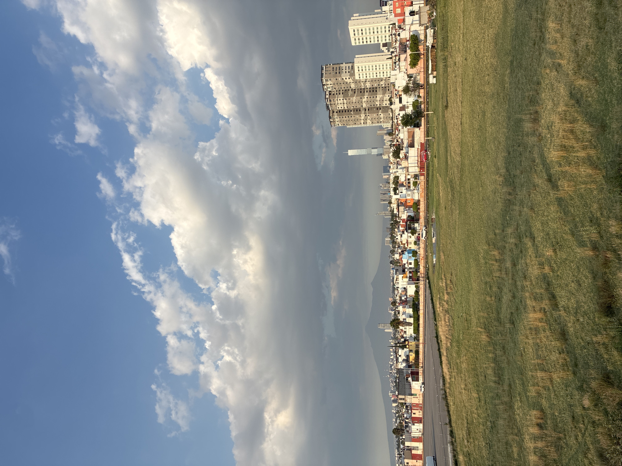 The image shows a view of San Bernardino Tlaxcalancingo, Mexico, on a day with varied weather conditions.
To the left, the sky is blue with white, puffy clouds. Further towards the middle of the image, the sky darkens, with heavier, darker clouds suggesting an approaching storm. In the distance, a mountain range is visible, obscured somewhat by the cloud cover.
The city itself is laid out in a grid pattern, with buildings of various sizes and colors. In the foreground, there is a road running parallel to a large grassy field. The city appears densely populated, with a mix of residential and commercial structures. Several tall apartment buildings are visible in the distance.