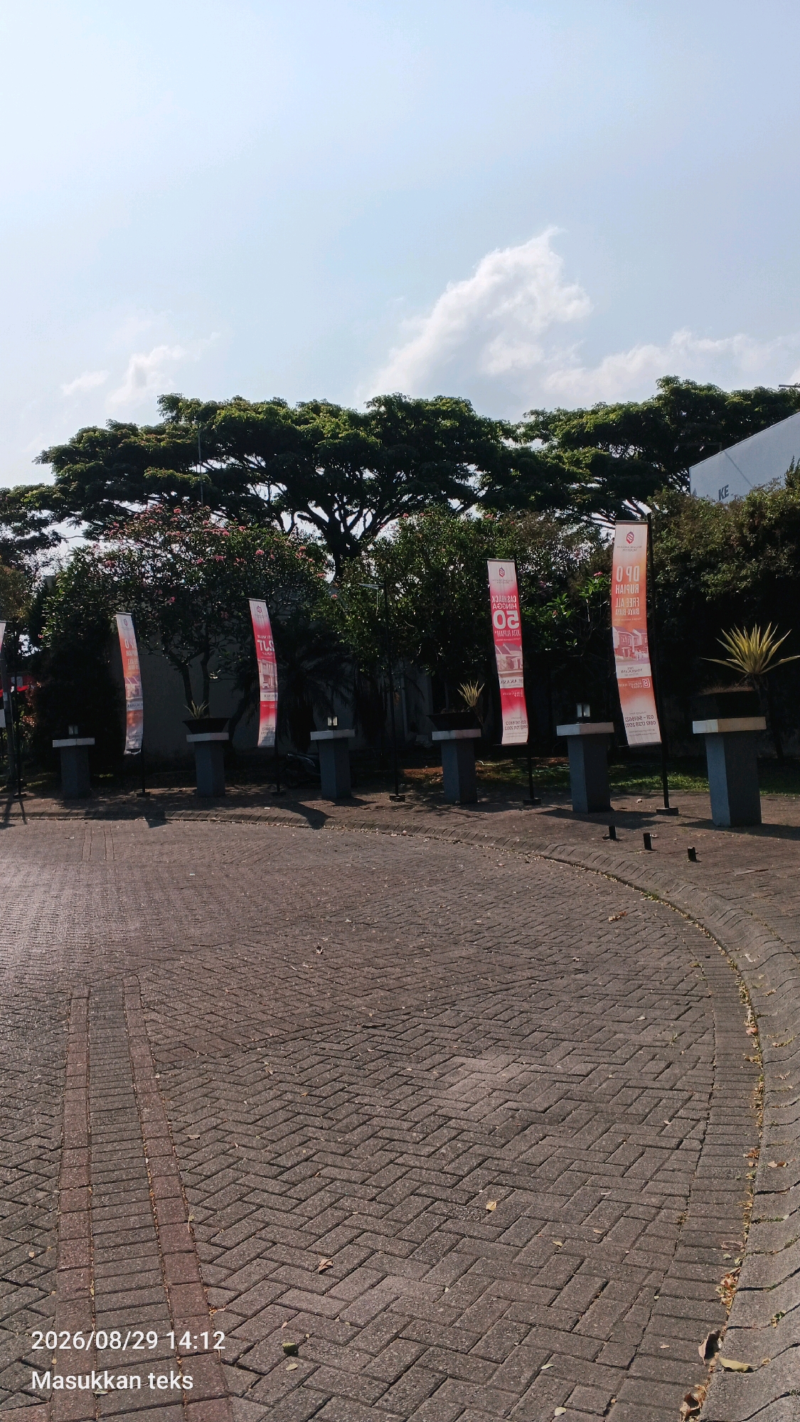 A paved walkway curves around a grassy area with several tall, rectangular banners standing upright. The banners are predominantly red and white, with some featuring text and graphics that suggest a promotional or informational purpose. Behind the banners, a dense line of trees forms a natural backdrop, with a particularly large, leafy tree dominating the upper portion of the frame. The sky above is bright blue with scattered white clouds. The walkway is made of interlocking brick pavers laid in a herringbone pattern. The timestamp "2026/08/29 14:12" and the text "Masukkan teks" are overlaid on the lower left of the image. The scene appears to be an outdoor public space, possibly near a building, given the presence of the banners and paved area.