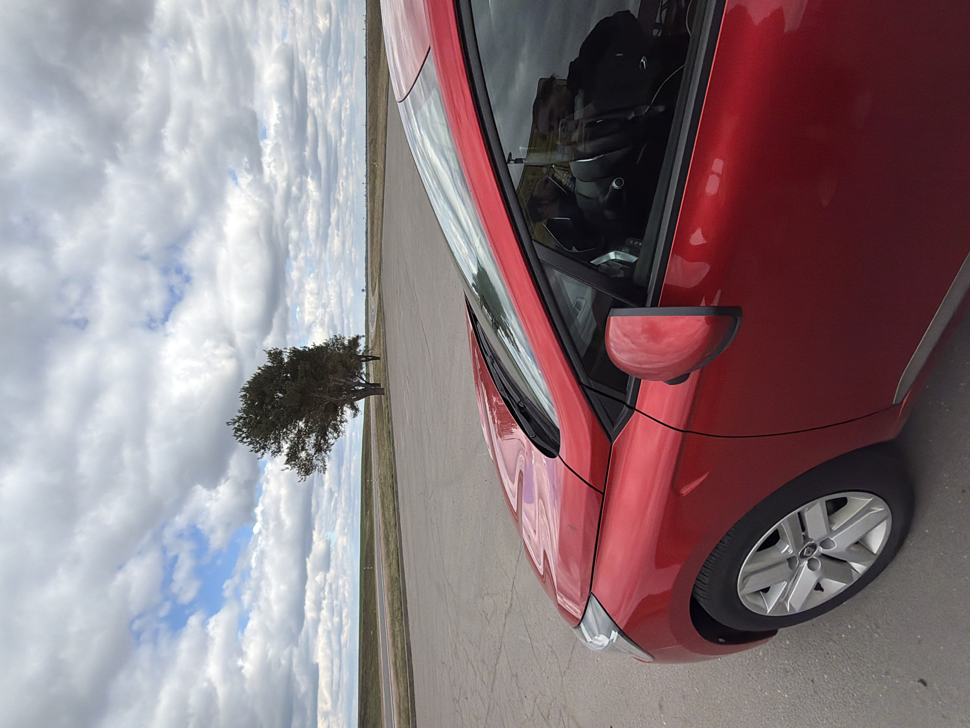 A close-up, angled shot captures a red car parked on an expansive paved surface, possibly an airfield or large parking area. The sky is heavily clouded with patches of blue visible. A single, mature tree stands at a distance across the tarmac. The environment appears to be in a rural location potentially within the Абухаўскі сельскі Савет area of Belarus.
