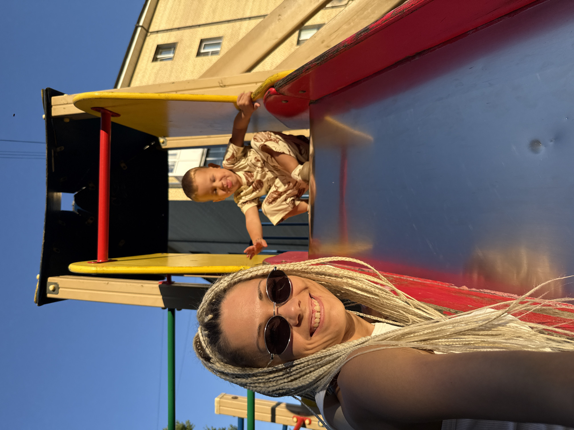 A woman with blonde braids and sunglasses is in the foreground of the image, smiling at the camera. Behind her, a young child is sitting on a slide, sticking out their tongue. The child is wearing a patterned outfit. They are on a playground with a blue sky and a building in the background. The scene appears to be set in Rusanovo, Russia.