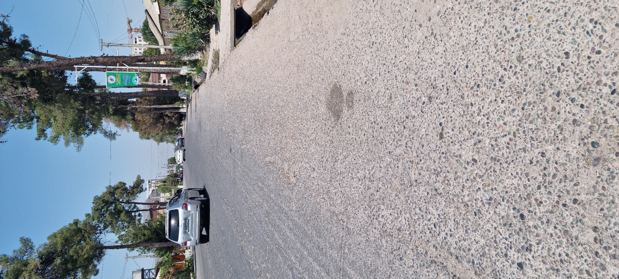 The image shows a street in Ismoili Somoni, Tajikistan. The street is paved with asphalt and concrete, and there are trees lining the sides of the road. A silver SUV is parked on the side of the road, and there are several other cars driving on the street. The sky is blue and clear, and the sun is shining. There is a drainage ditch on the right side of the road. There are also several buildings visible in the background, including houses and a construction crane. A green banner with text in an unfamiliar language is hanging from a lamppost.