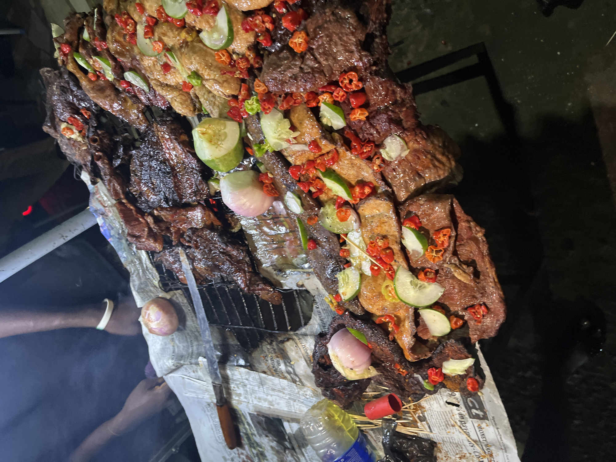 The image depicts an outdoor food stall in Abuja, Nigeria, likely a street-side eatery. A large, grilled assortment of meats, including what appears to be beef and chicken, is prominently displayed on a wire rack, covered with newspaper and garnished with sliced cucumbers, red chili peppers, and onions. The food is smoky, suggesting it has just been cooked or is still being prepared. In the background, two individuals are partially visible, their hands tending to the food or the stall. One person wears a white bracelet. A bottle of cooking oil and a red cylindrical object, possibly a container for spices or condiments, are also present. The lighting suggests it is nighttime or a dimly lit area.
