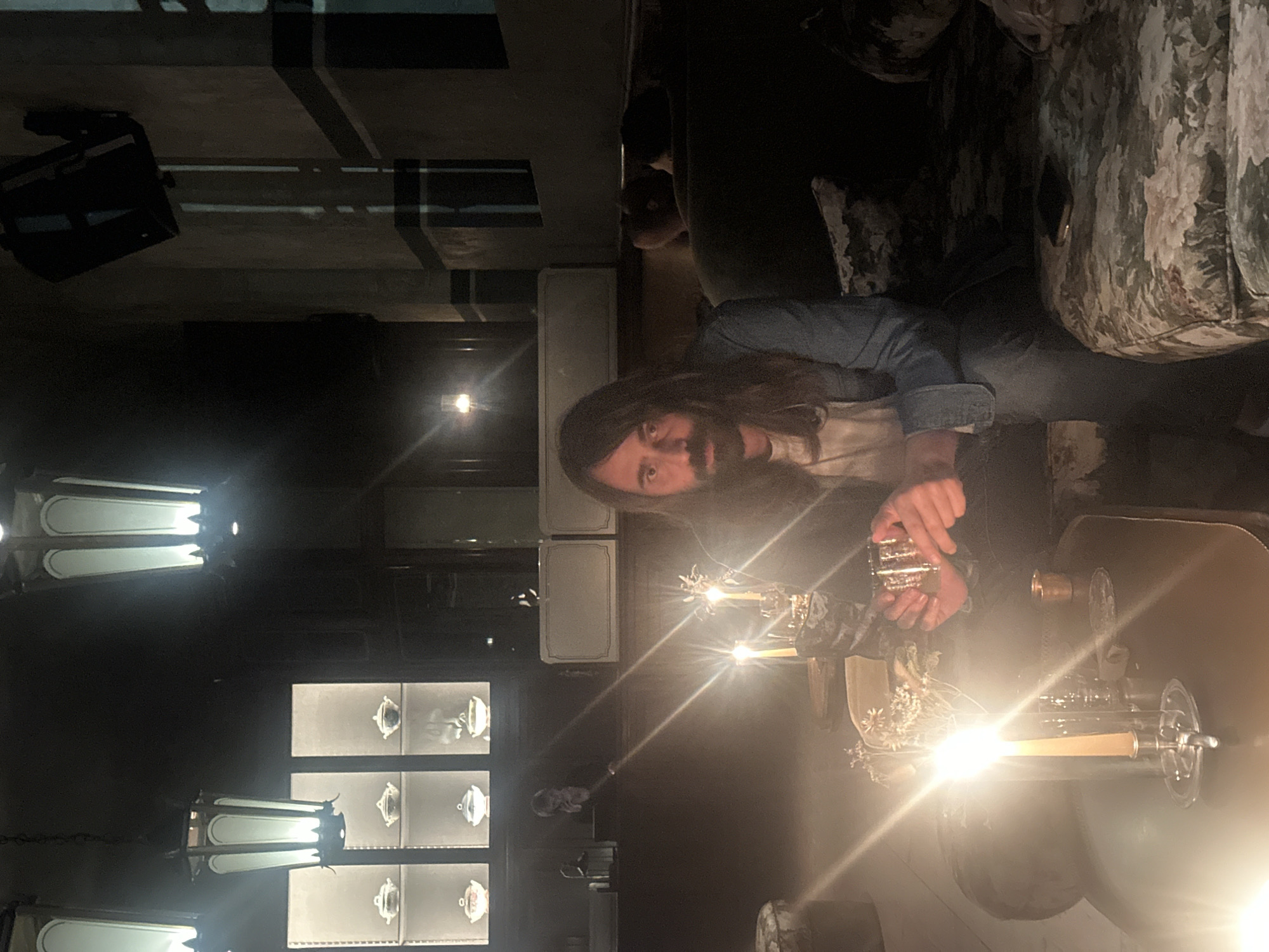 A man with long hair and a beard is holding a small, ornamented glass and sitting next to an illuminated bar, possibly at a restaurant or bar. He is wearing a denim jacket over a light-colored shirt. The bar features decorative elements, including what appears to be a candelabra. In the background, there is a display cabinet with decorative objects. The overall lighting in the room is dim, with the bar lighting providing a central focal point. The location is most likely in Milan, Italy.
