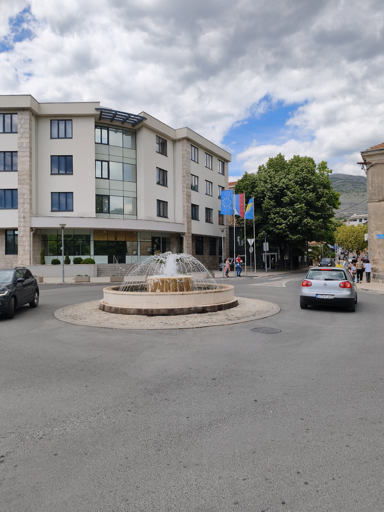 The image shows a street scene in Trebinje, Bosnia and Herzegovina. A roundabout with a fountain is the central feature. A light-colored, multi-story building stands beside the roundabout. Three flags (European Union, Croatia, and Bosnia and Herzegovina) are displayed near the building. A dark-colored car is exiting the roundabout on the left, and a silver Volkswagen is exiting to the right. Several people are walking on the sidewalk on the right side of the road. The sky is cloudy with patches of blue. A mountain is visible in the background on the right side.