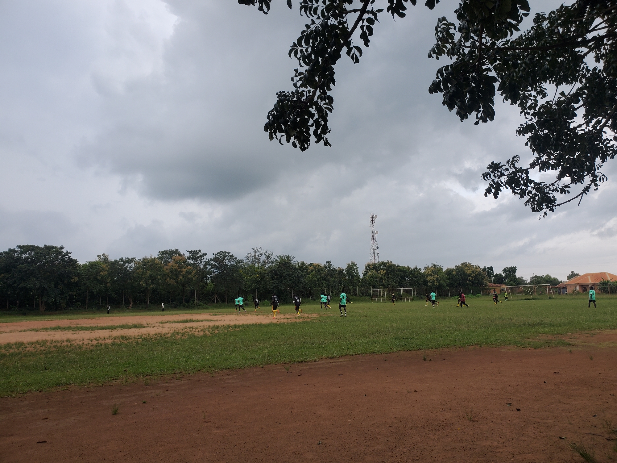 An aerial perspective reveals a football match in progress on a field under a cloudy sky. The field, a mix of dirt and grass, is bordered by trees and a few buildings in the distance. Several players, clad in green and black jerseys, are actively engaged in the game. A mobile tower stands tall in the background, and a soccer goal can be seen on the right side of the field. The overall scene suggests an outdoor sports activity in a rural or semi-urban setting in Miango, Nigeria.