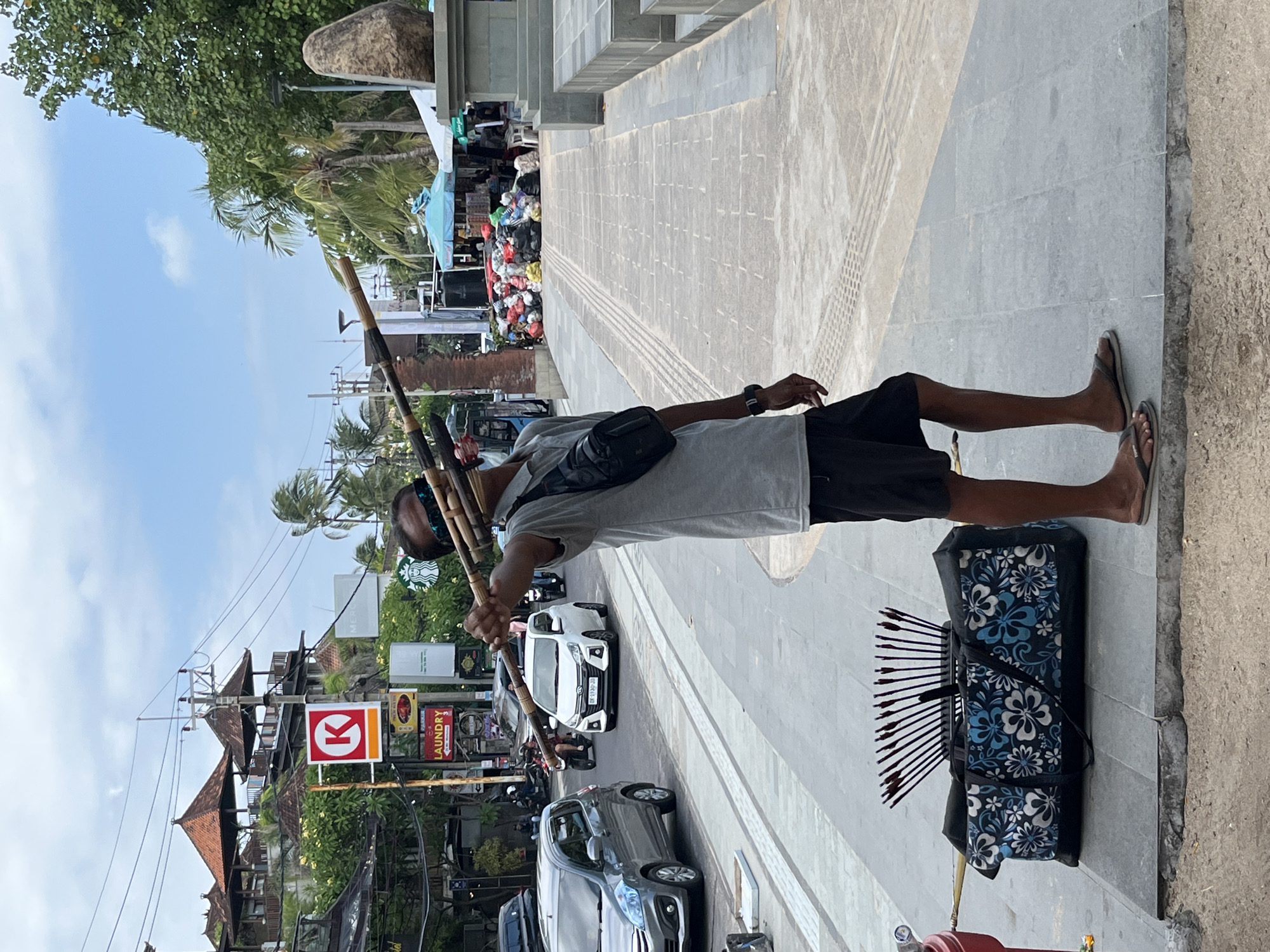 A person stands on a paved sidewalk, holding several long, thin bamboo poles vertically. They are wearing a gray t-shirt, black shorts, and flip-flops, with a black bag slung across their chest. Next to them on the ground is a black bag with a blue and white floral pattern, from which a fan of arrows emerges. The person appears to be a vendor selling archery equipment.
The scene is a street in Kuta, Indonesia, with shops and signs visible in the background. Several vehicles, including cars and motorcycles, are on the road. The sky is blue with some clouds, suggesting it is daytime, likely morning or afternoon, with fair weather. The architecture of the buildings, with their tiled roofs, and the presence of palm trees indicate a tropical location. A red and white circular sign with a "K" is visible on one of the buildings, possibly indicating a business or local establishment. A "Laundry" sign is also visible. The overall impression is a bustling street scene in a tourist area.