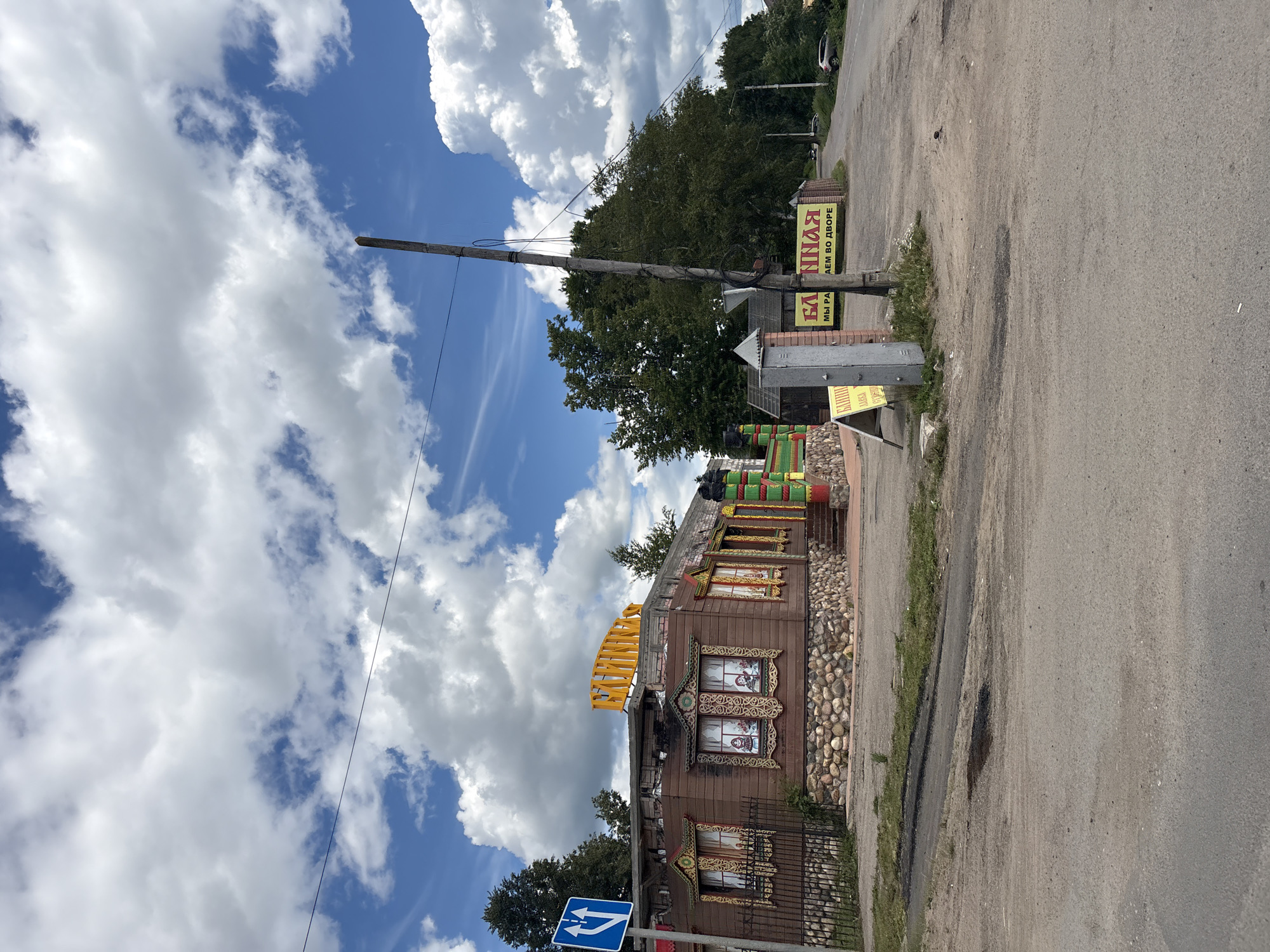 The image captures a scene in Pereslavl-Zalessky, Russia, on a bright, partly cloudy day. The sky is a vibrant blue with large, fluffy white clouds. A wooden pole with wires stretches across the frame, suggesting the presence of electricity or communication lines.
In the foreground, a distinctive wooden building stands out. It is adorned with colorful, ornate window frames and a stone foundation. The building's architecture has a traditional Russian feel, with intricate carvings and decorative elements. A bright yellow structure, possibly a sign or part of the building's facade, is visible on the roof. A black metal fence runs along the front of the building.
To the right of the building, a paved road is visible, with patches of grass and some minor damage to the asphalt. There are also several signs with Russian text. One prominent yellow sign reads "БАННАЯ" (BATHHOUSE) with "ЕМ ВО ДВОРЕ" (IN THE YARD) below it. Another sign, partially visible, seems to have "МЫ РА" on it. Further down the road, a traffic sign with blue arrows indicates a direction. Trees and foliage line the background, and a car can be seen in the distance on the road. The overall impression is a sunny day in a semi-rural or suburban setting, with a focus on a unique, decorated building.