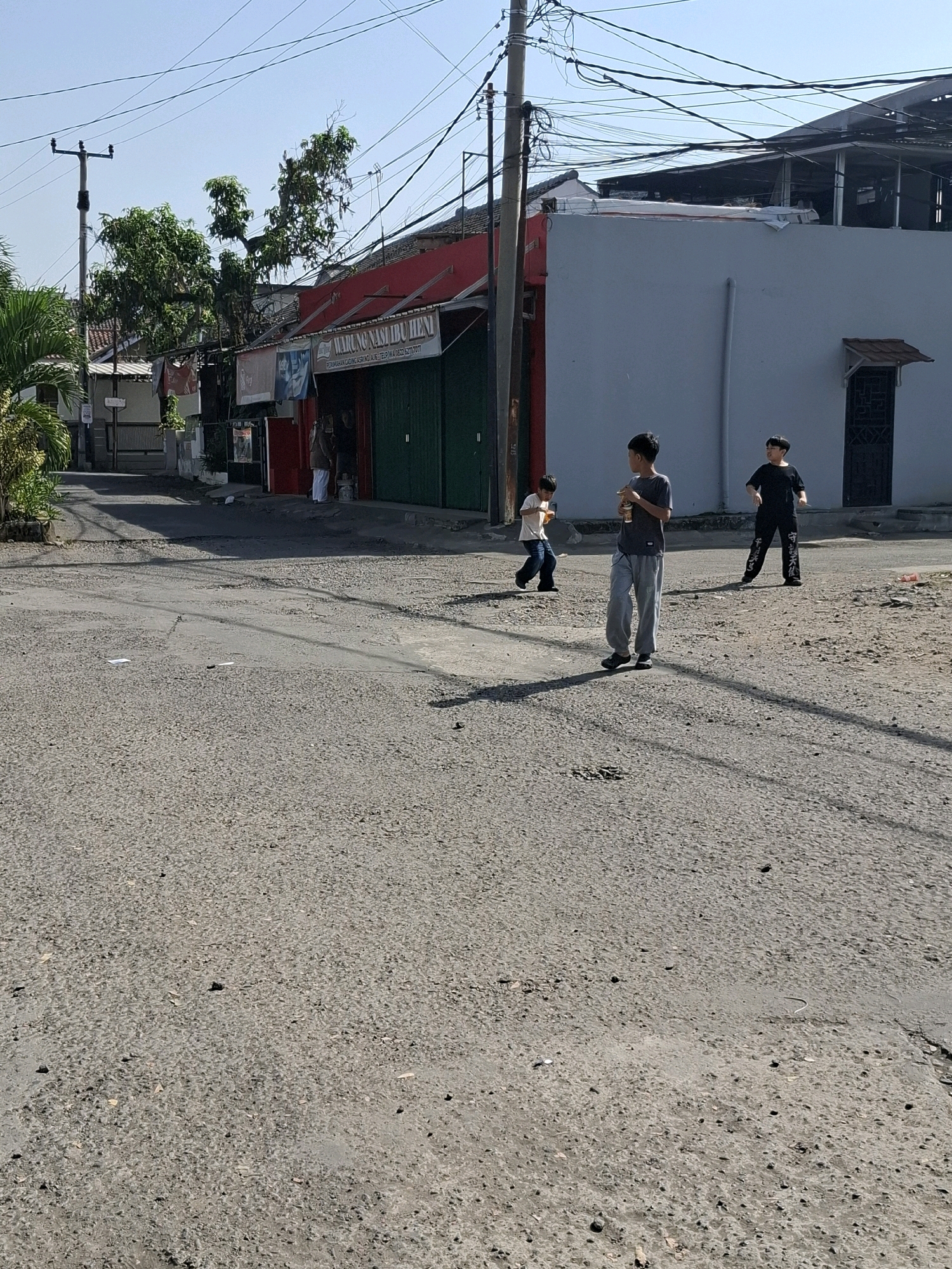 The image captures a street scene in Karangtengah, Indonesia, under a clear blue sky. Three young boys are present. One boy, wearing a dark t-shirt and black pants with white lettering, stands on the right side of the street, looking towards the left. A second boy, wearing a gray t-shirt and light gray sweatpants, is in the center, holding a yellow cylindrical object and looking to the left. The third boy, in a light t-shirt and dark jeans, is on the left, also facing left and holding a yellow object. The street is paved with asphalt and shows signs of wear, with some potholes. Buildings line the right side of the street, including a structure with a light blue wall and a red-fronted building with green doors and a sign that reads "Warung Nasi Ibu Heni." Overhead, a network of power lines crisscrosses the sky, supported by utility poles. Greenery, including palm fronds and trees, is visible on the left side of the image. The overall impression is a casual, everyday scene in a residential or mixed-use urban area.
