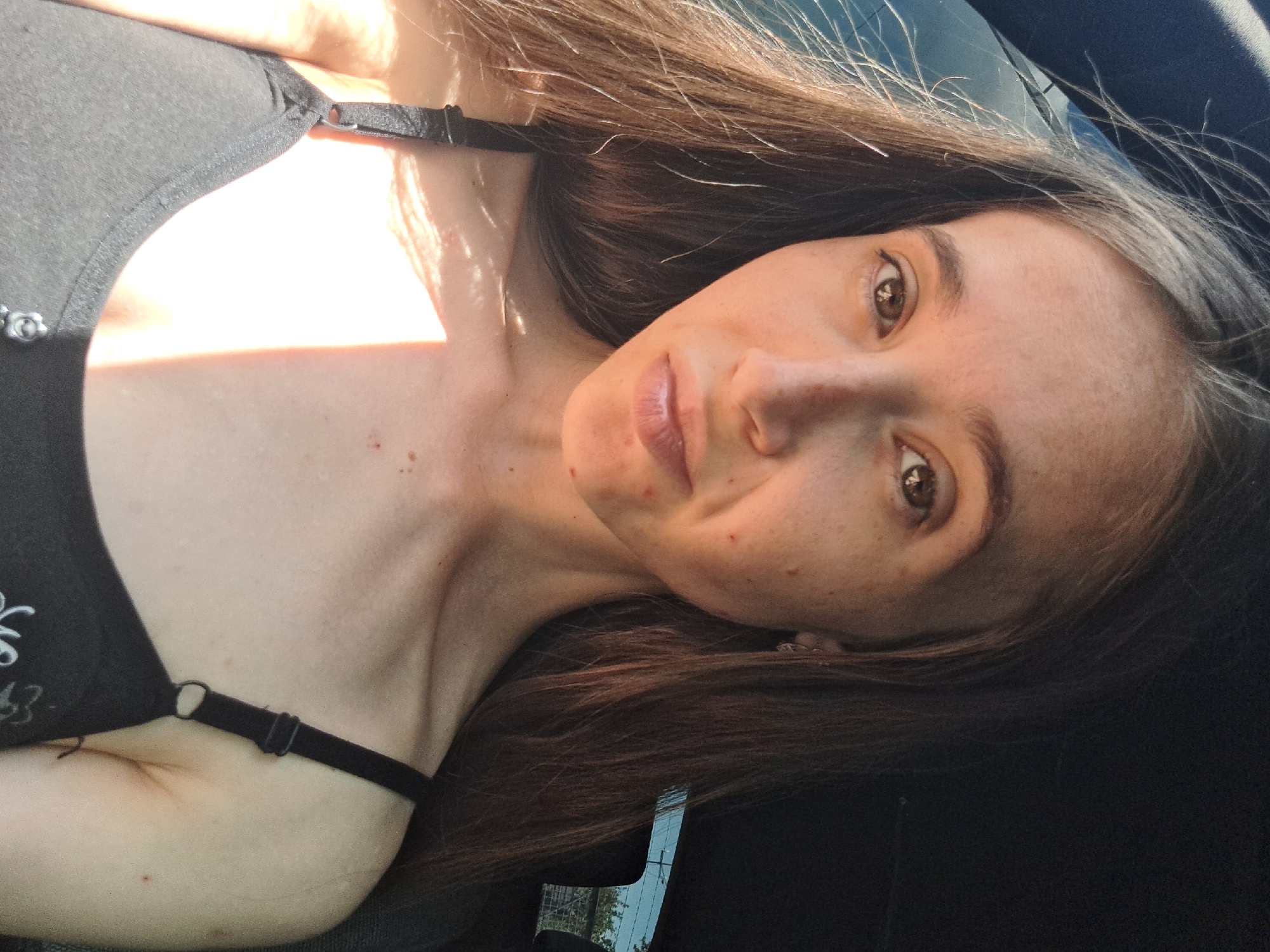 A young woman with long, dark brown hair and brown eyes is pictured in a close-up, front-facing shot. She is wearing a black tank top. Her skin has freckles and some blemishes. Sunlight is casting a bright streak across her left shoulder and chest. The background is dark, suggesting she is inside a vehicle, with a hint of the outdoors visible through a window on the right side. The location is Vypasne, Ukraine.