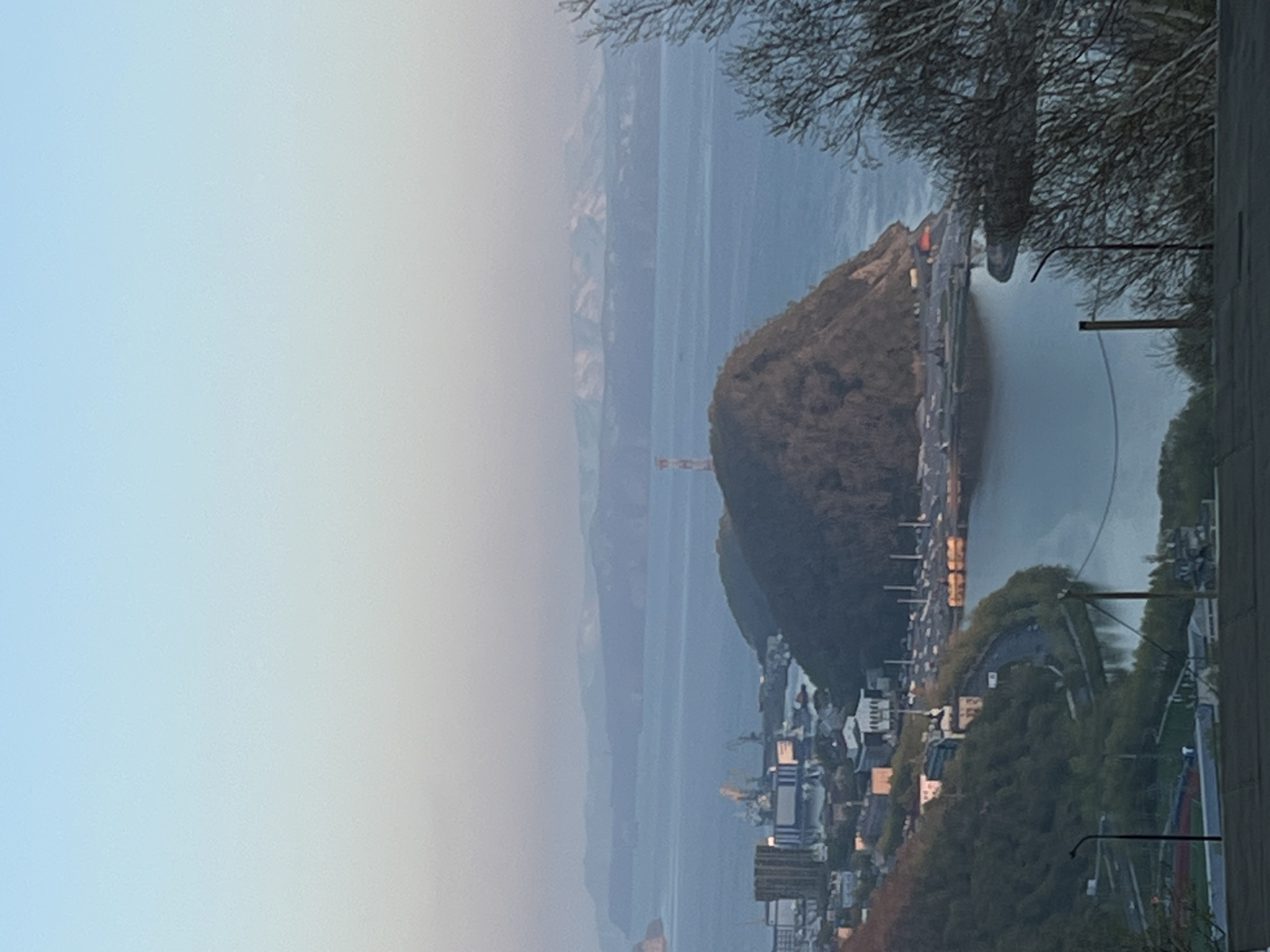 The image displays a broad, slightly hazy panoramic view of a coastal city, identified as Petropavlovsk-Kamchatsky, Russia, framed by a soft, clear sky. The overall lighting suggests either early morning or late afternoon.
In the foreground, the lower right section features the bare branches of a tree, indicating a colder or dormant season. Adjacent to this, parts of a building with a light-colored wall and utility poles or wires are visible.
The midground is dominated by a large, dark, tree-covered hill or small mountain that projects into a body of water, likely a bay or ocean. A road or highway with faint vehicle lights winds around the base of this hill. To the left of the hill, the urban area of the city spreads along the coastline, comprising numerous buildings and what appear to be industrial or port structures. A tall, slender communications tower stands prominently on a distant part of the hill or shoreline.
Beyond the expansive body of water, a long chain of layered mountains stretches across the horizon. Their peaks are softened by atmospheric haze, blending into the pale blue sky. The sky itself transitions from a gentle light blue at the top to an almost white hue closer to the distant mountains, indicative of atmospheric conditions.
No specific human activity is clearly depicted, but the urban setting implies ongoing daily life. There is no visible text in the image. The scene evokes a sense of tranquility and highlights the dramatic natural environment surrounding the city.