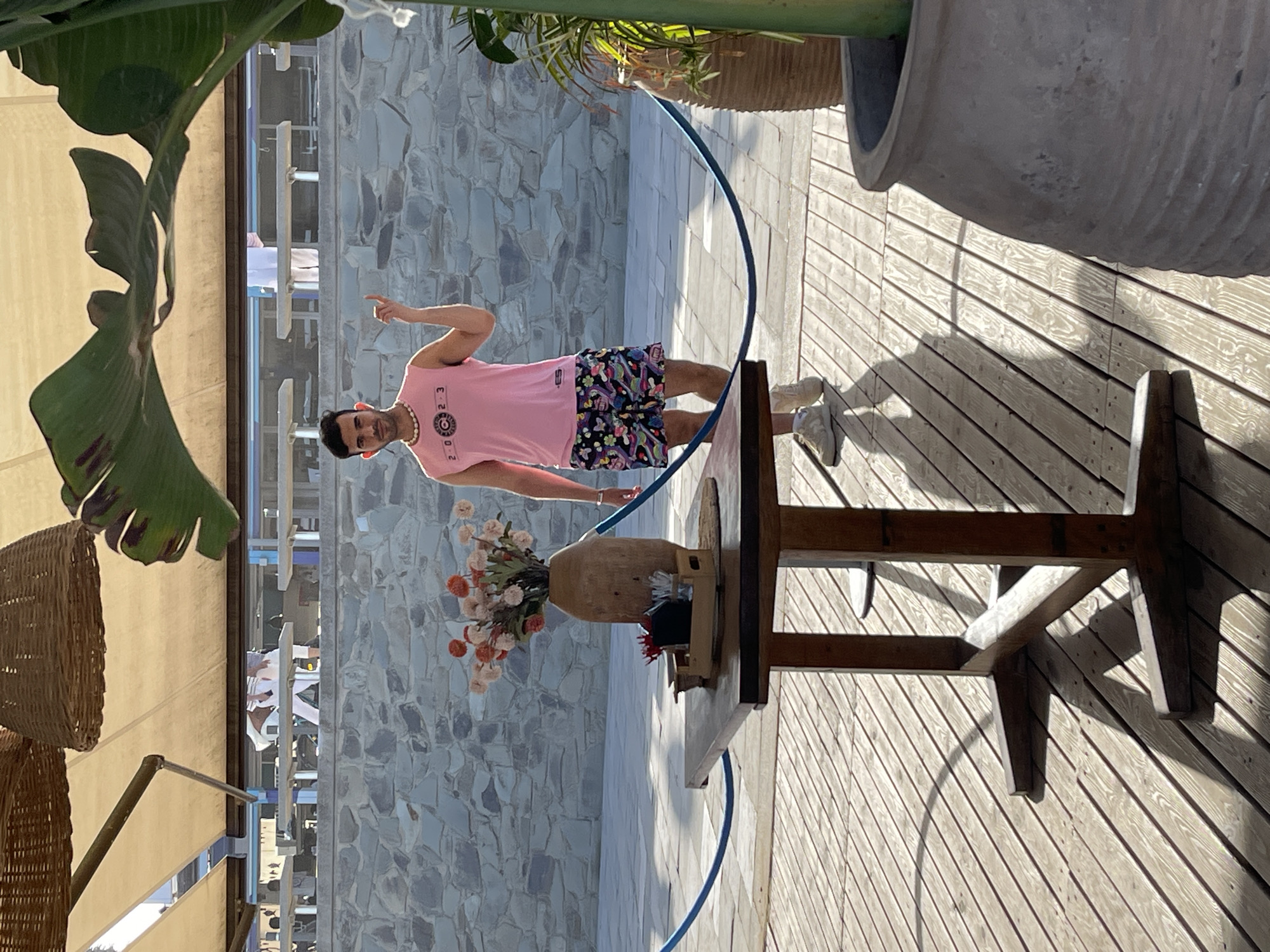 A man in a pink t-shirt and floral shorts stands on a wooden deck, pointing to his left. He wears a pearl necklace and white sneakers. In front of him, a wooden table holds a vase of flowers and a small black object. The scene is outdoors, with a stone wall in the background and large green leaves from a plant in the foreground. A blue hose curves across the deck. Sunlight casts shadows on the wooden planks, suggesting it is daytime. The man's expression is neutral. The setting appears to be a relaxed outdoor space, possibly a patio or garden area, with elements suggestive of a warm climate.