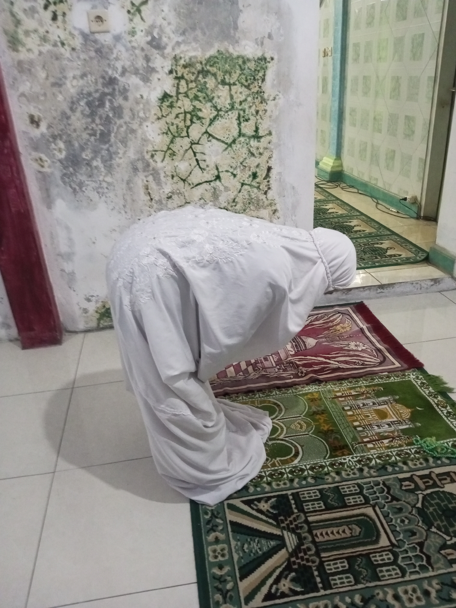 A person in a white prayer garment is performing Salat (Islamic prayer) on a prayer rug in what appears to be a room with tiled floors. The person is in a bowing position, with their head lowered towards the rug. The prayer rug has intricate patterns in red, green, and white. In the background, there's a wall with signs of mold or water damage, and some patterned tiling on another wall. Electrical outlets are visible on the walls. The scene appears to be indoors.