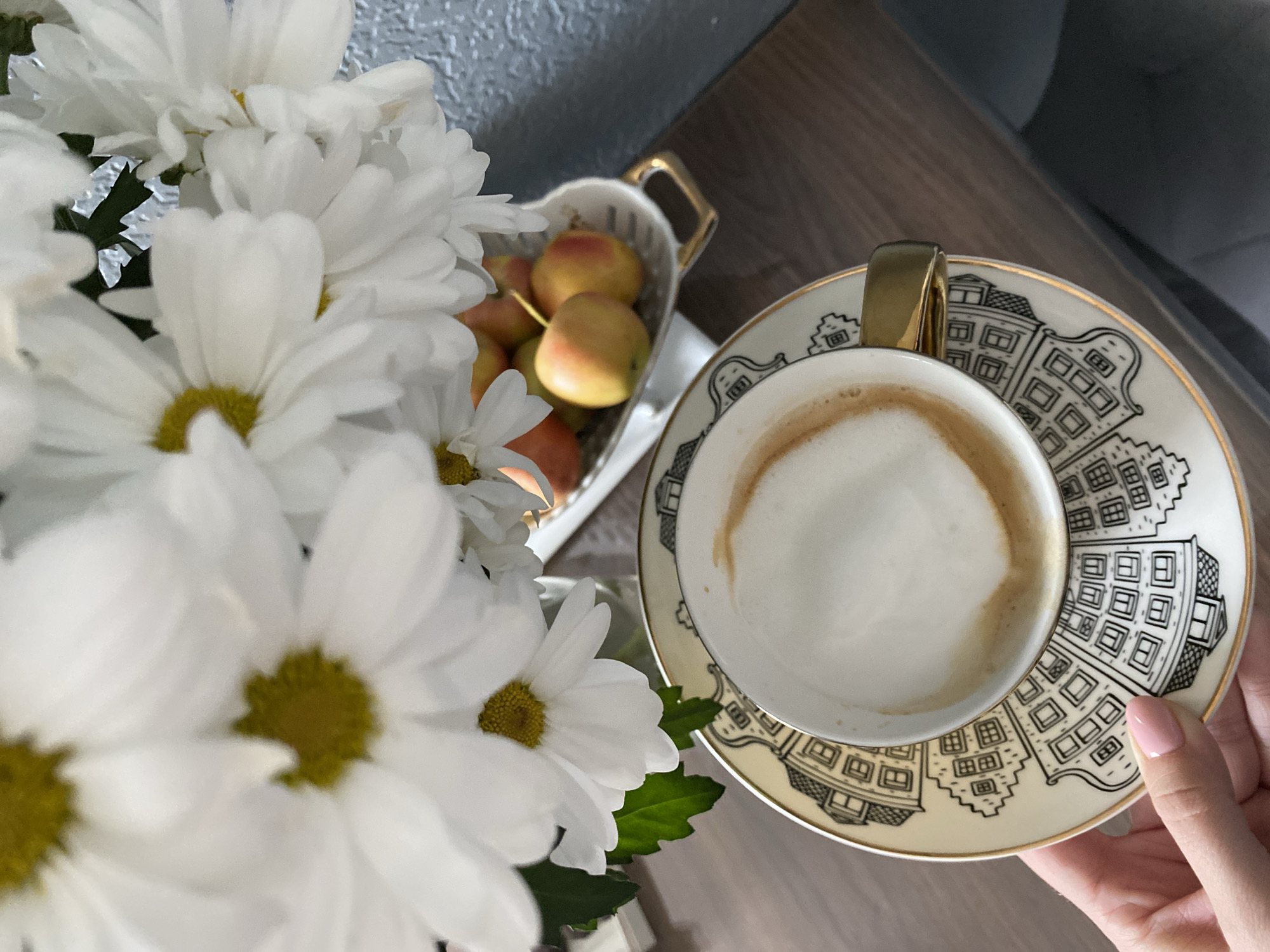 An overhead, slightly angled shot captures a serene indoor scene, likely in Saint Petersburg, Russia. The central focus is a white ceramic coffee cup with a golden handle, brimming with a creamy latte or cappuccino, complete with a foam top and a faint ring of coffee. A person's hand, with light pink manicured nails, gently cradles the matching saucer beneath the cup. This saucer is a notable feature, adorned with a detailed black and white illustration depicting European-style city buildings or houses, and finished with a delicate golden rim.
To the left, a prominent bouquet of white chrysanthemum-like flowers with bright yellow centers fills the foreground, slightly out of focus, lending a fresh and vibrant touch to the composition. Behind the flowers, a charming heart-shaped white ceramic dish, also with golden handles, holds a cluster of small, round fruits that resemble reddish-yellow crabapples or small apples. The scene is set on a warm, brown wooden surface, with a textured blue-grey wall visible in the background. The natural lighting suggests it is daytime. The overall ambiance evokes a sense of calm and enjoyment, characteristic of a pleasant coffee break or a relaxing moment in a home or cafe. There is no visible text in the image.
