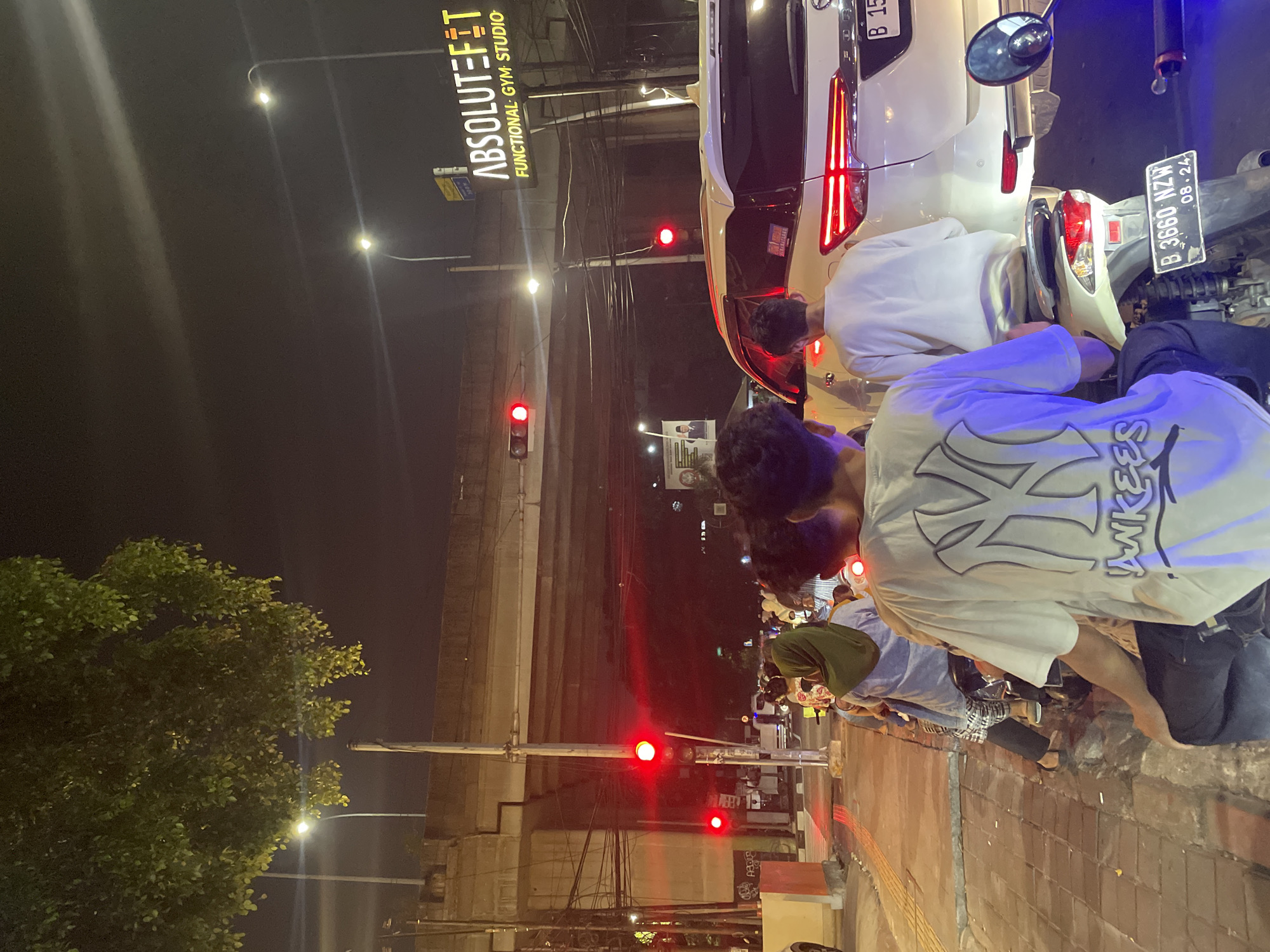 A nighttime scene captures a group of people gathered around a white car and a scooter on a street in North Jakarta, Indonesia. The subjects appear to be young men, with one wearing a white t-shirt featuring a graphic of a "NY" logo and the word "YANKEES" across the front. Another person is visible in a light blue shirt, and someone else is wearing a green headscarf.
The setting is urban, characterized by streetlights, traffic lights displaying red, power lines, and a textured brick sidewalk. A large, leafy green tree is prominent on the left side of the image. A neon sign with the text "ABSOLUTE FIT FUNCTIONAL GYM STUDIO" is visible in the upper portion of the frame. License plates are visible on the vehicles, including "B 15" on the car and "B 3660 NZW 08.24" on the scooter.
The activity is unclear, but the people seem to be standing around, possibly socializing or engaged in an informal gathering. The time of day is clearly night, indicated by the artificial lighting and the dark sky. The atmosphere appears casual and possibly social, with no overt signs of distress or urgency.