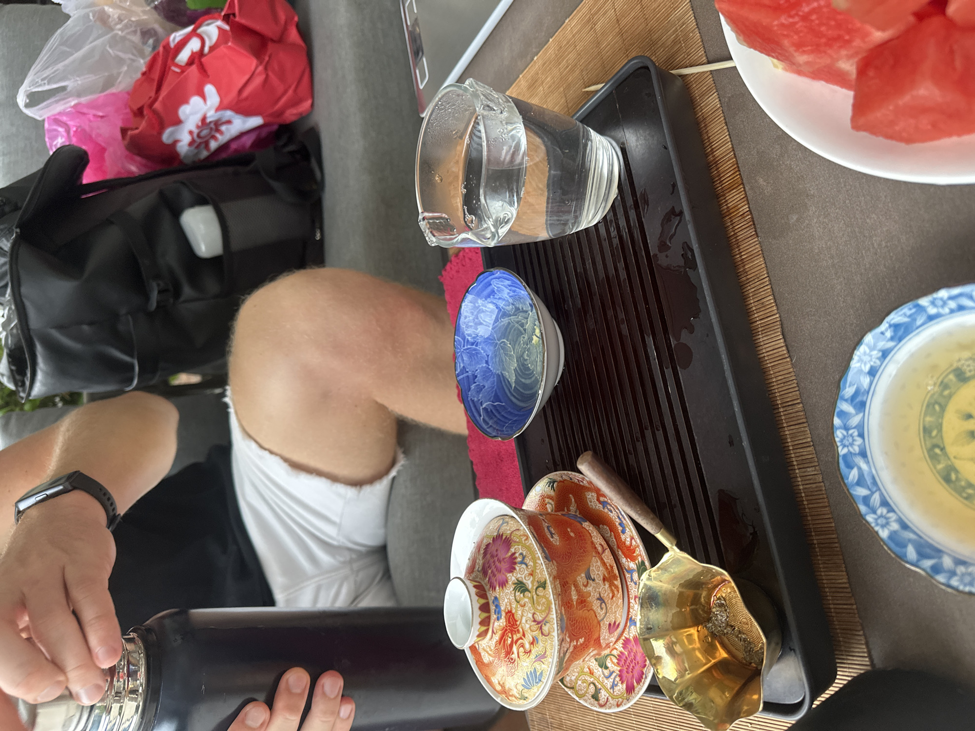 In an outdoor setting, possibly a patio, a person is preparing tea. A person's hands are visible, holding the lid of a large black thermos. They are wearing a black t-shirt and white shorts, and a black fitness tracker is on their left wrist. A tea set is laid out on a dark rectangular tray placed on a bamboo mat. The tea set includes a lidded teacup with intricate orange and gold dragon and floral designs, a saucer, a small blue and white bowl with a floral pattern, and a golden ladle containing tea leaves. A clear glass pitcher with a pour spout holds clear liquid. In the background, a large black backpack and several colorful plastic bags are visible on a grey upholstered surface, likely a couch. A bowl of cut watermelon is in the lower right corner of the image. The scene suggests a moment of relaxation or a tea ceremony.