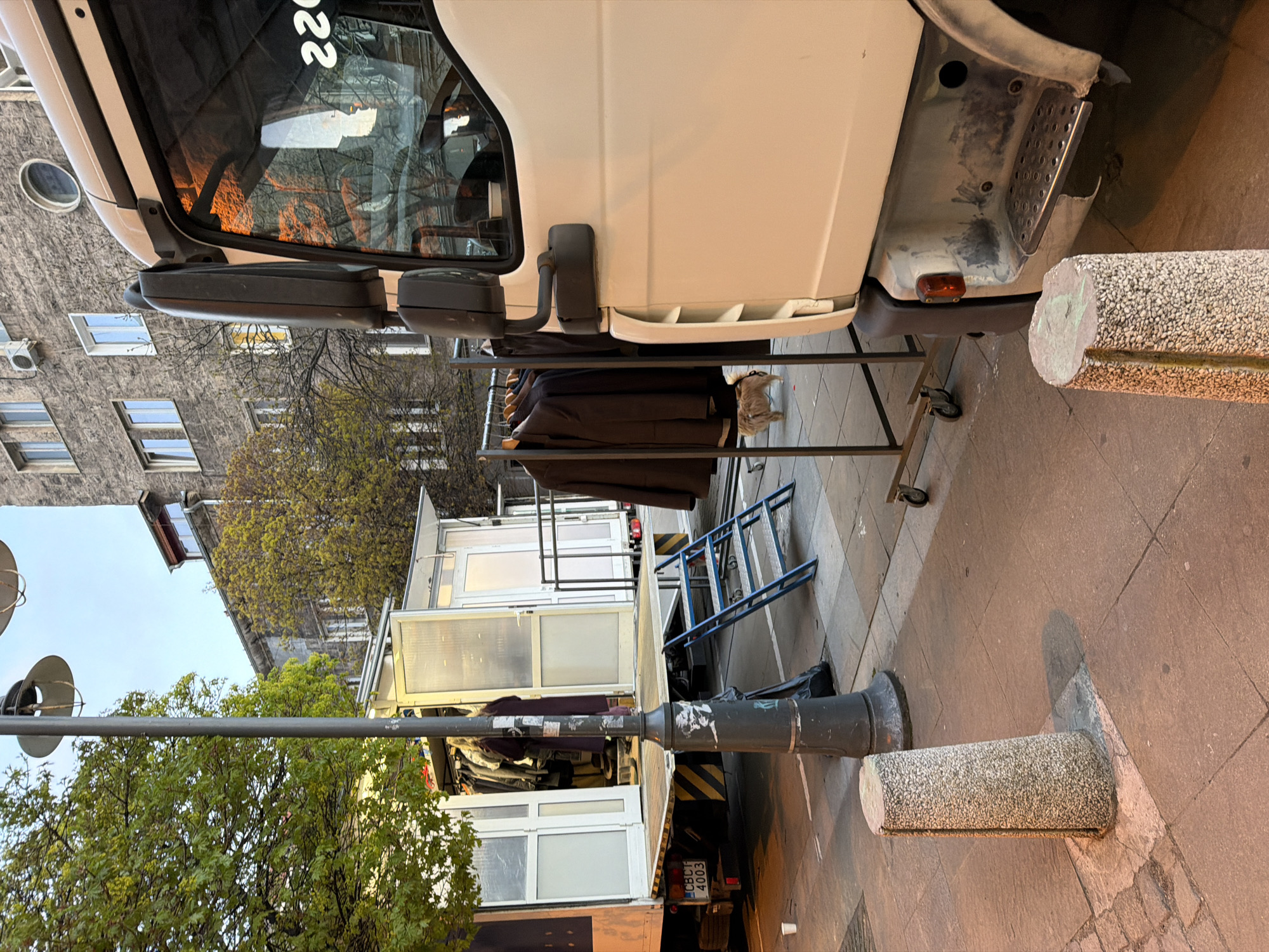 The image shows a parked truck in an urban environment, likely a street in Unknown, Unknown. The truck's cab is white, with a visible side mirror and part of the front bumper. A metal clothes rack, holding what appear to be dark-colored clothes, stands beside the truck. Behind the truck, there is a white structure with transparent windows, which appears to be a mobile retail space or display unit. A blue ladder leans against the unit. The scene is framed by a light pole on the left and a cylindrical bollard on the right. In the background, there's a multi-story building with windows and trees. The lighting suggests either early morning or late afternoon.