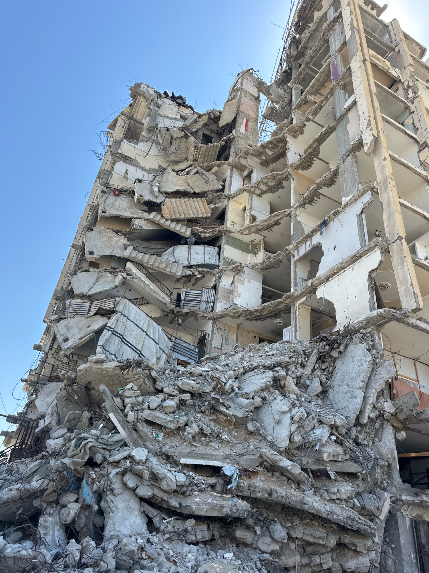 This image depicts the severe structural damage to a multi-story building in the city of Gaza, Palestinian Territories. The building has been largely destroyed, with its facade heavily compromised and many floors collapsed. Large sections of concrete, rebar, and other building materials are strewn across the ground in a large pile of rubble at the base of the structure. The interior of the building is exposed, revealing broken staircases, remaining wall sections, and hollowed-out rooms. The sky above is clear and blue, providing a stark contrast to the devastation below. There are no people visible in the image, and the scene appears to be one of aftermath and destruction.