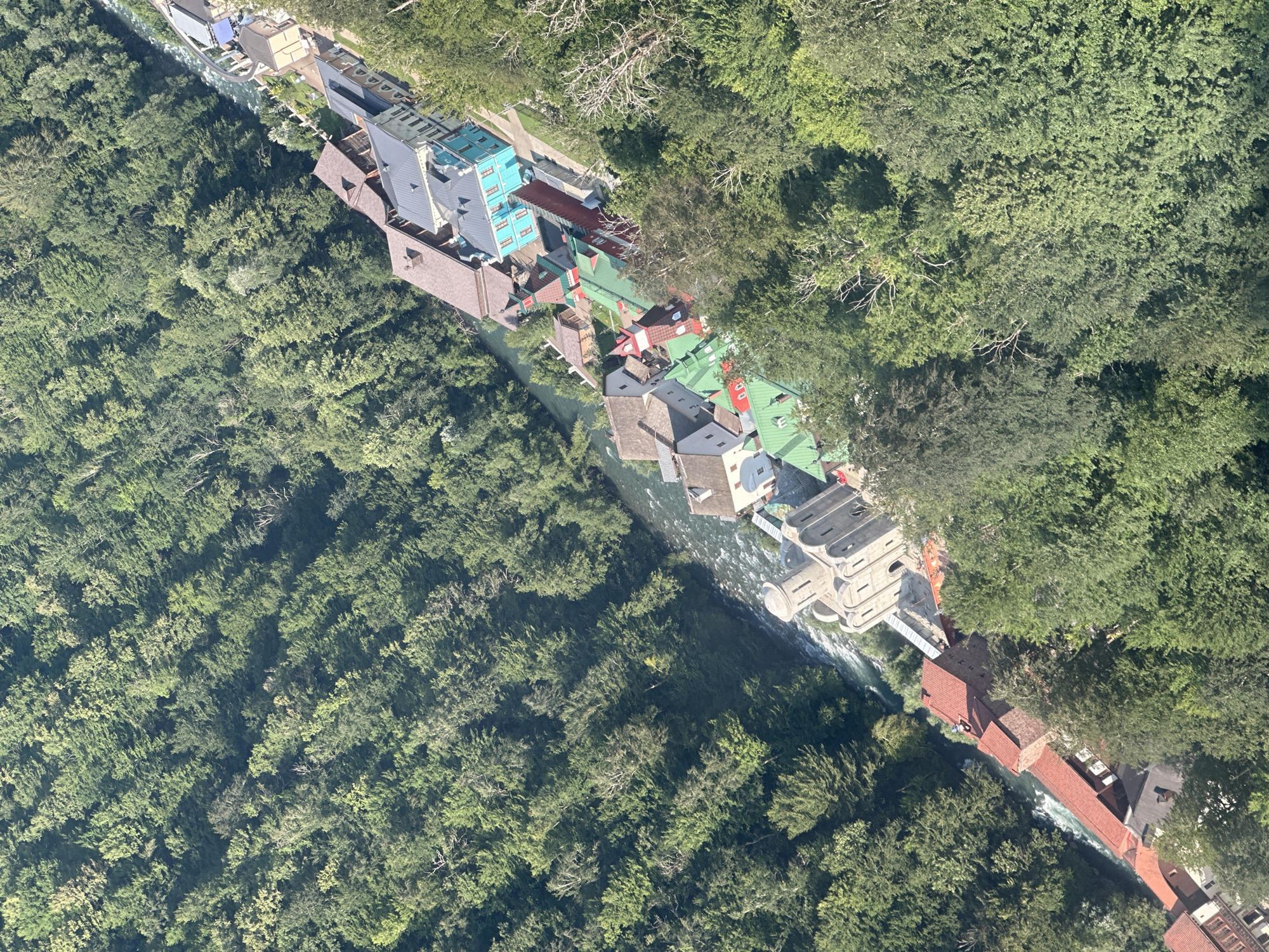 The aerial image shows a village nestled within a lush green forest, with a river flowing through it. The village features a collection of buildings with colorful roofs, including red, green, and blue. Some buildings have a traditional architectural style, while others appear more modern. The river appears to be fast-flowing, with white water rapids visible in several areas. The surrounding forest is dense with a variety of trees, creating a verdant landscape. The village appears to be situated on a slope, with the river cutting through a valley. The scene suggests a remote and picturesque location, likely in a mountainous region. Based on the context provided, this location is identified as Estosadok, Russia.