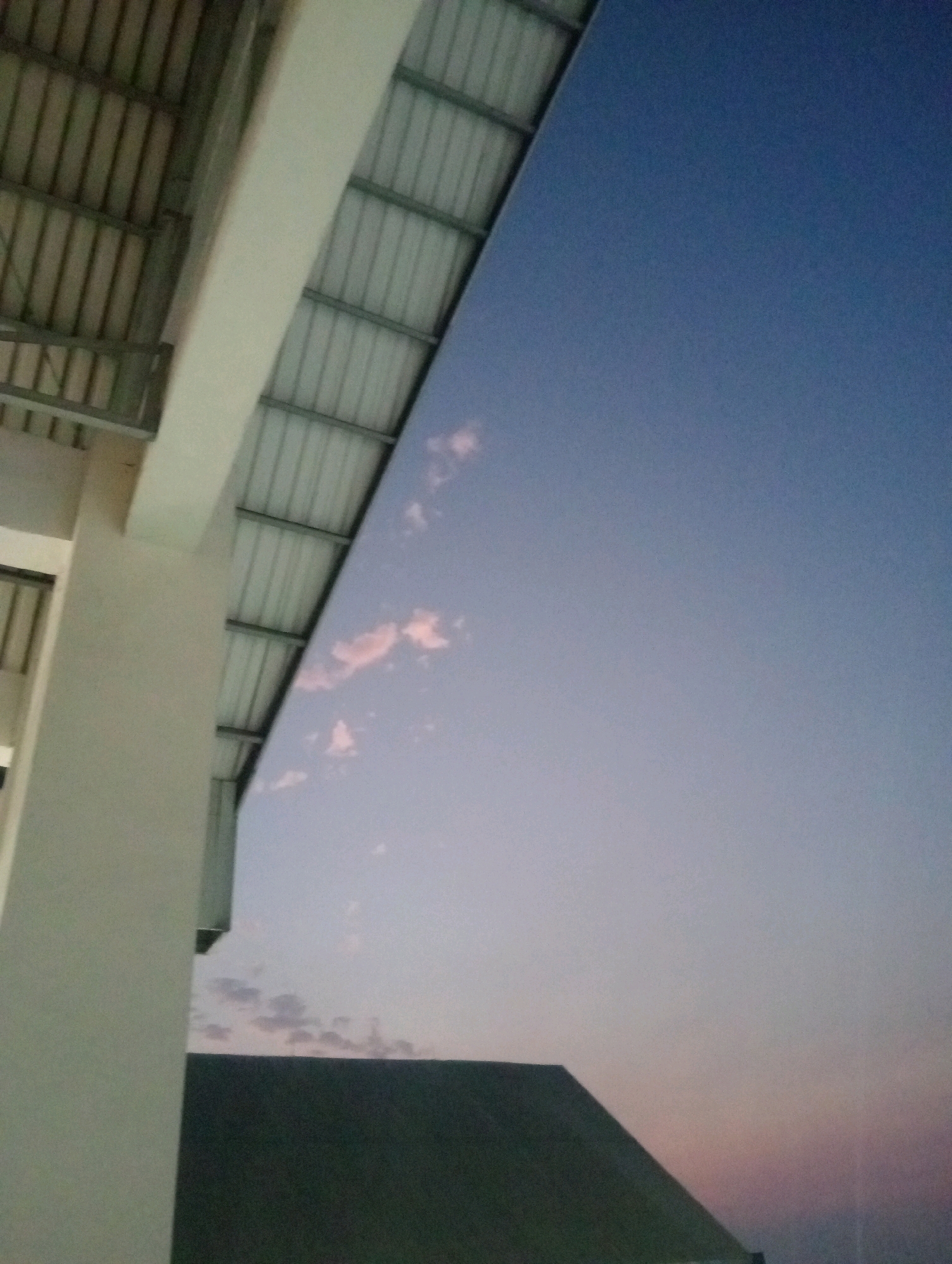 The image depicts a perspective looking upwards towards the sky, with the edge of a building's corrugated metal roof in the foreground on the left. The roof is supported by a light-colored pillar. The sky is a gradient of deep blue at the top, fading to a lighter, almost lavender hue towards the horizon, indicating it is either dawn or dusk. Scattered, wispy pink clouds are visible against the lighter parts of the sky. In the lower portion of the image, the dark, angled roof of another building is visible, forming a stark contrast with the sky. The scene appears to be an outdoor setting, possibly a residential or commercial area, with the architecture suggesting a modern construction. The lighting conditions suggest low light, likely early morning or late evening. There are no people or animals visible in the image. The overall atmosphere is serene and quiet, with the focus being on the architectural elements and the sky. The location is Batu, Indonesia.
