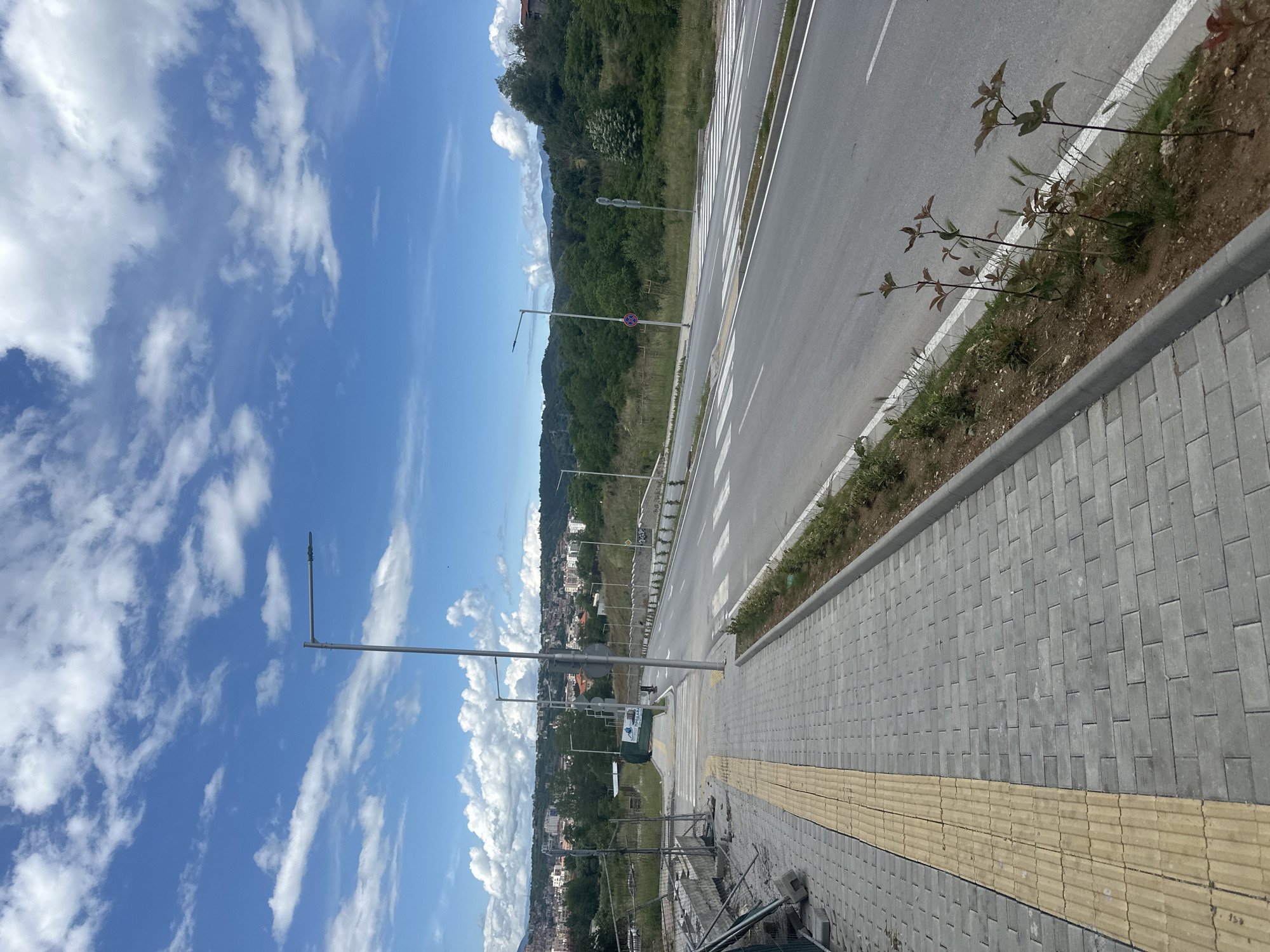 The image presents a bright, sunny daytime scene in Blagoevgrad, Bulgaria, capturing a road and an adjacent pedestrian sidewalk. The sky is a vivid blue, dotted with scattered white cumulus clouds.
On the right side of the frame, an asphalt road with multiple lanes is visible, marked by distinct white lines, including a prominent pedestrian zebra crossing. Adjacent to the road, a well-maintained sidewalk is paved with grey rectangular blocks, featuring a continuous yellow tactile paving strip designed for visually impaired individuals.
To the left of the road, a strip of green grass and small, recently planted saplings separates the infrastructure from the natural landscape. In the background, rolling green hills and mountains, densely covered with trees, stretch across the horizon. Beneath these hills and extending into the middle ground, a townscape with numerous buildings can be seen. Several tall, modern streetlights with extended arms illuminate the road and sidewalk area.
There are no people or specific activities depicted in the image, suggesting a tranquil moment. Notable details include a red circular sign with a white horizontal bar, indicating "No Entry" or "Do Not Enter," attached to a streetlight pole. Below it, a white sign with green Cyrillic text is partially visible, which appears to indicate a "Construction" zone ("СТРОЕЖ" or "СТРОИТЕЛЕН"), corresponding with some visible construction fencing in the lower left foreground. The scene combines modern urban infrastructure with a picturesque natural environment.