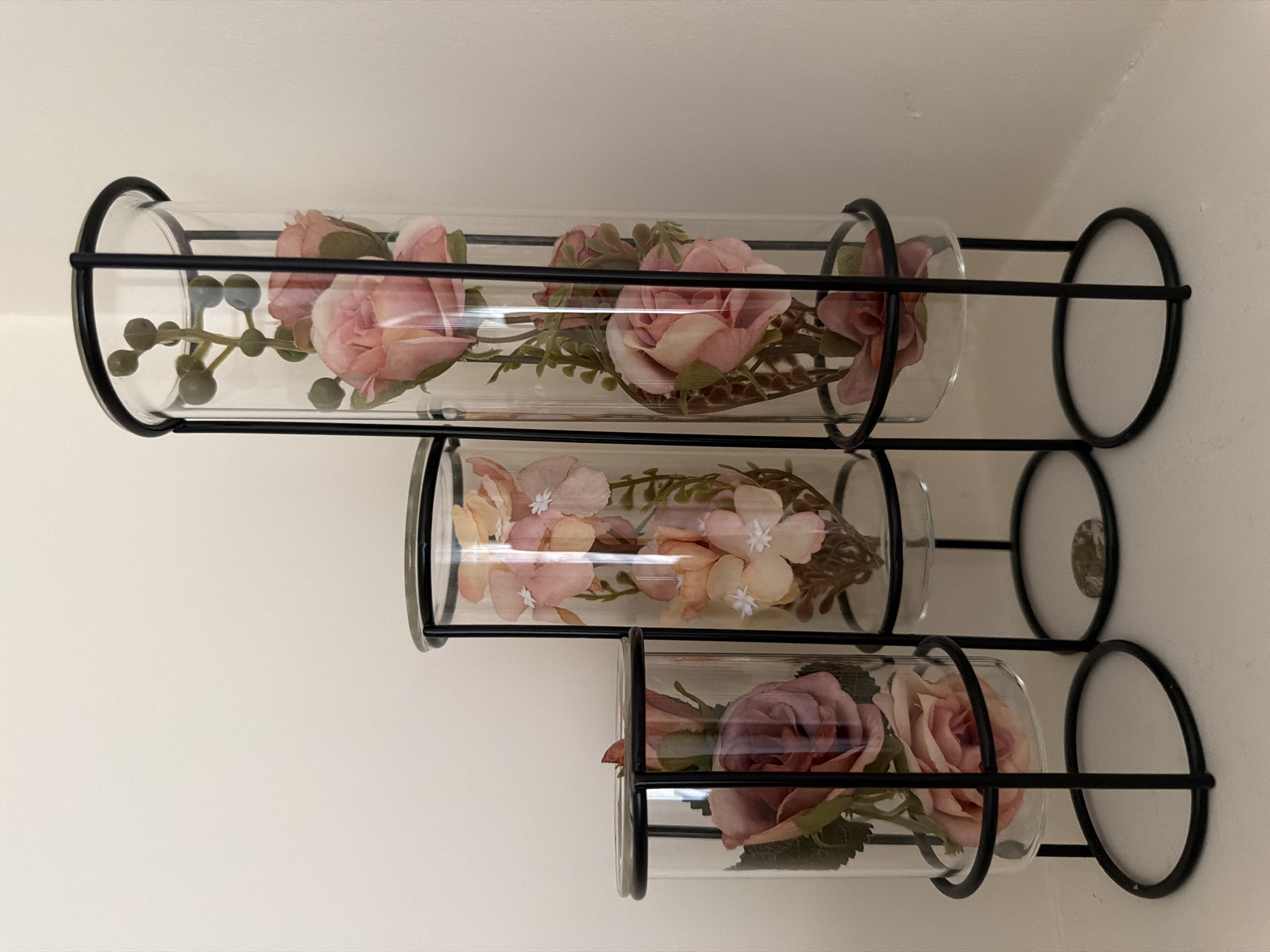 Three glass cylindrical vases containing artificial flowers are arranged on a shelf against a plain, light-colored wall. The vases are supported by black metal stands, with each vase varying in height and diameter, creating a tiered display.
The smallest vase on the left holds two dusty rose-colored roses and some green foliage. The middle vase, slightly taller and wider, contains several pale pink and peach-colored flowers with white centers, along with greenery. The tallest and widest vase on the right is filled with pale pink roses and a stem with small green berries.
The objects are illuminated by soft, ambient light, casting subtle shadows. The background is a simple, unadorned wall, suggesting an indoor setting. There are no people or animals present in the image. The scene appears to be a static, decorative arrangement.