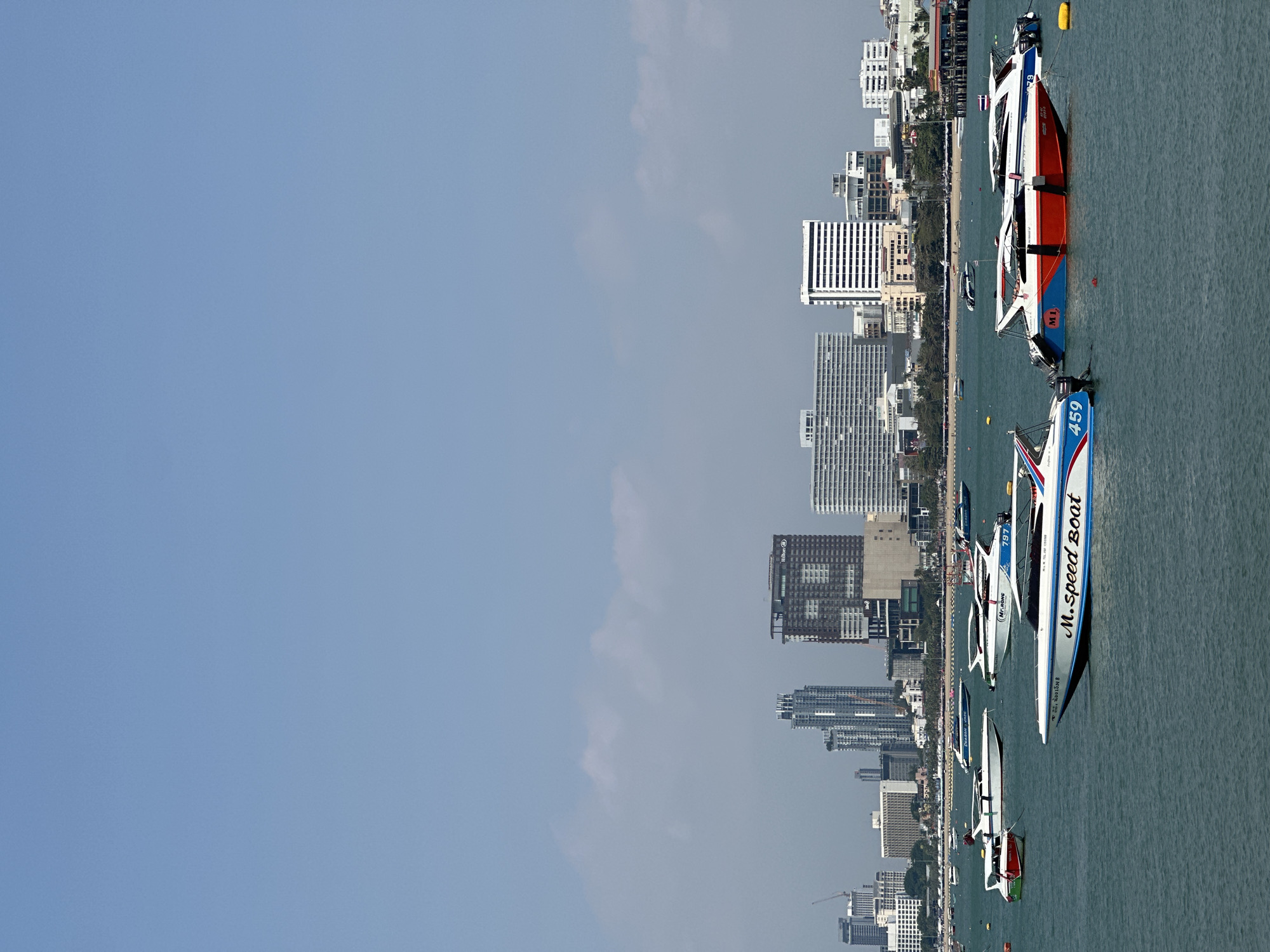 The image depicts a waterfront view of Pattaya City, Thailand. In the foreground, several speedboats are visible in the water. The city skyline features a mix of high-rise buildings and smaller structures, suggesting a coastal urban area. The sky is partly cloudy. No people are clearly visible in the image. One boat is labeled "M.Speed Boat" with the number 459. Another boat has the number 197.