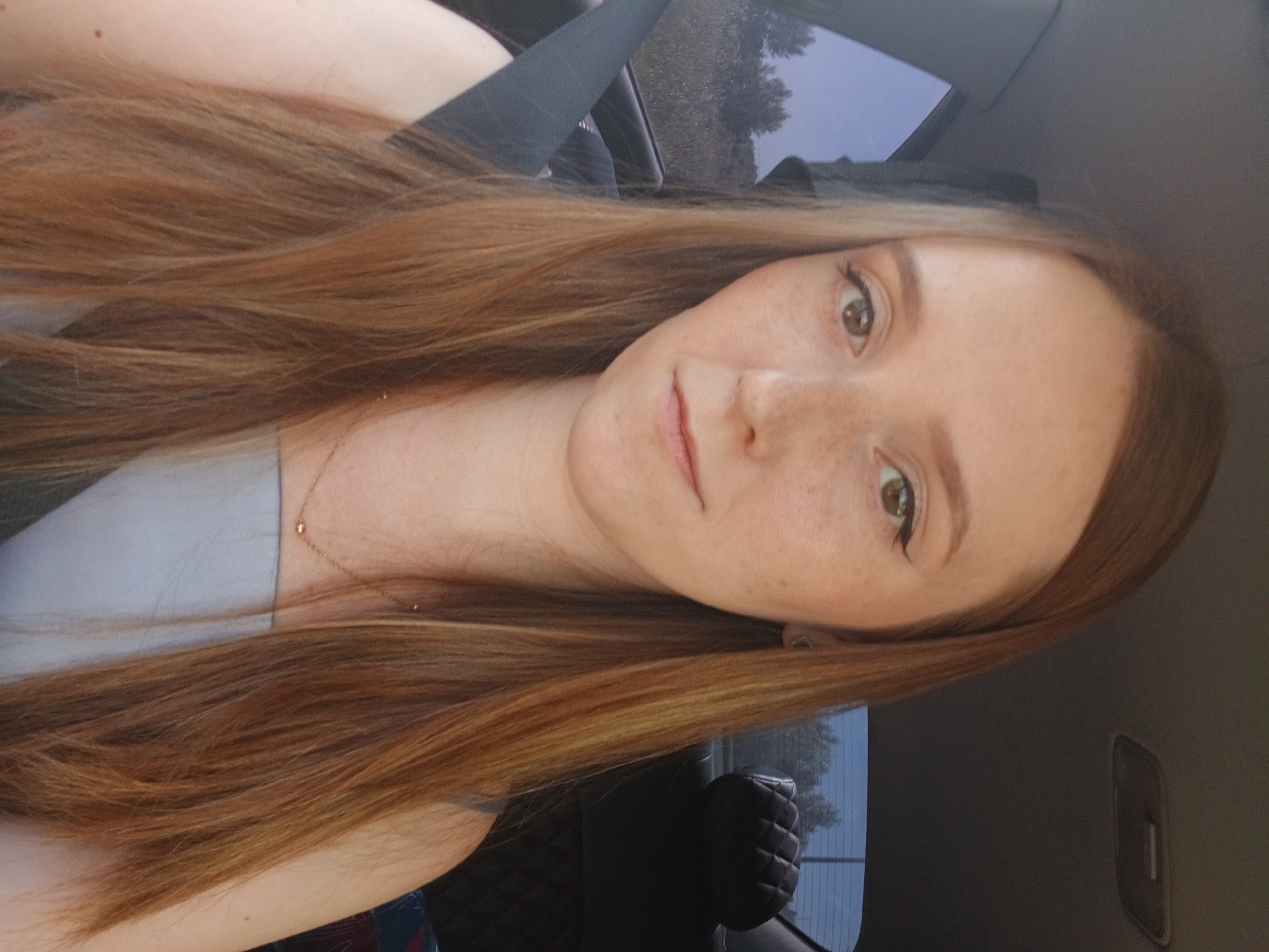 A medium shot of a young woman with long, auburn hair and freckles is taken from inside a car. She is wearing a light blue, strapless top and a thin gold necklace. Her hair falls around her shoulders, and she is looking directly at the camera. The background shows the interior of a car, including a seatbelt, car interior ceiling, and a quilted headrest. Through the car windows, trees and foliage are visible, suggesting a rural or suburban setting. The lighting suggests it is daytime.