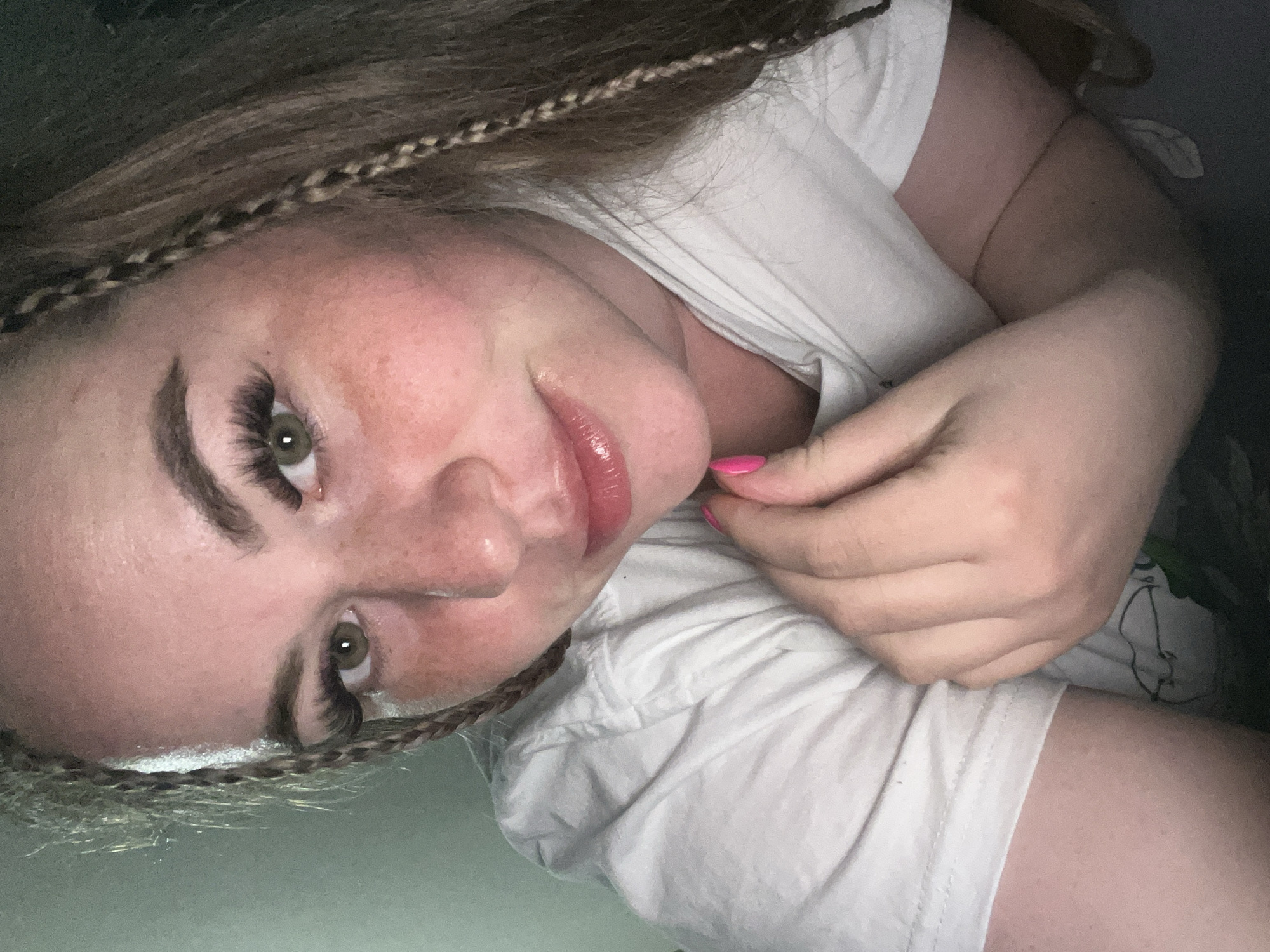 A close-up selfie of a fair-skinned woman with light brown hair featuring thin braids near her face, wearing a white t-shirt. Her face is tilted towards the camera, showcasing prominent eyelashes, defined eyebrows, and glossy lips. Her right hand is positioned near her chin with bright pink nail polish visible on one fingernail. The background is out of focus, revealing dark shades. The location cannot be verified based on image evidence.