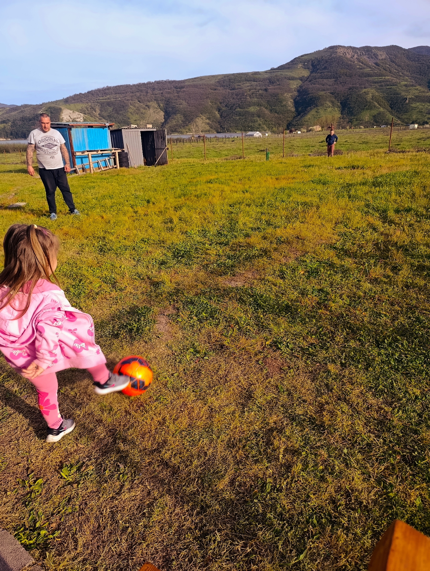 The image shows a young girl in pink sportswear kicking an orange and black soccer ball on a grassy field in what appears to be the city of Shiroko Pole, Bulgaria. A man wearing a grey t-shirt and dark pants stands to the left of the frame, near a blue and grey structure that may be small buildings or sheds. Another man stands in the distance. A mountain range is visible in the background. The lighting suggests it is daytime, possibly late afternoon.