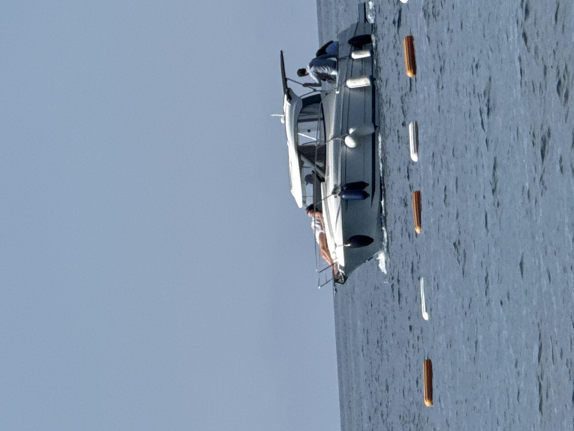 A vertical shot depicts a motorboat on the sea, with a clear blue sky in the background. The motorboat, mostly white, has multiple passengers, including one person visible on a ladder near the water. The sea is a moderate blue with slight ripples visible. Floating near the boat are several buoys, alternating between white and orange. The location is likely near Budva, Montenegro, given its coastal setting and presence of boats.