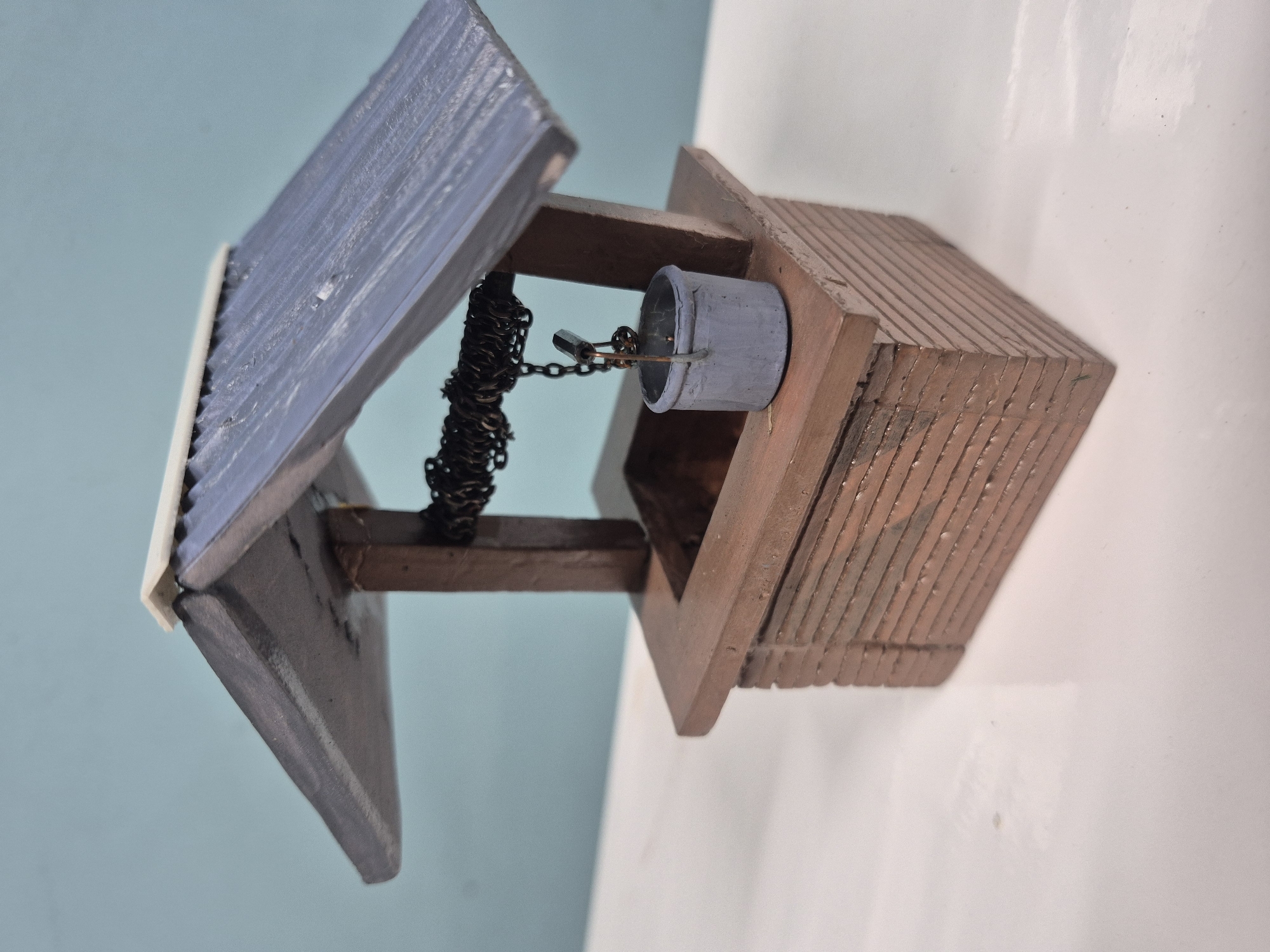 The image shows a miniature water well model with a light blue and white background on the left and a plain white background on the right. The well structure is mostly brown, with a small gray bucket hanging from a black chain. The well's roof, angled to the left, is blue with a white trim. The structure itself appears to be made of stacked wood pieces. There is no information to confirm the presence of the location of Chaikovsky, Russia.
