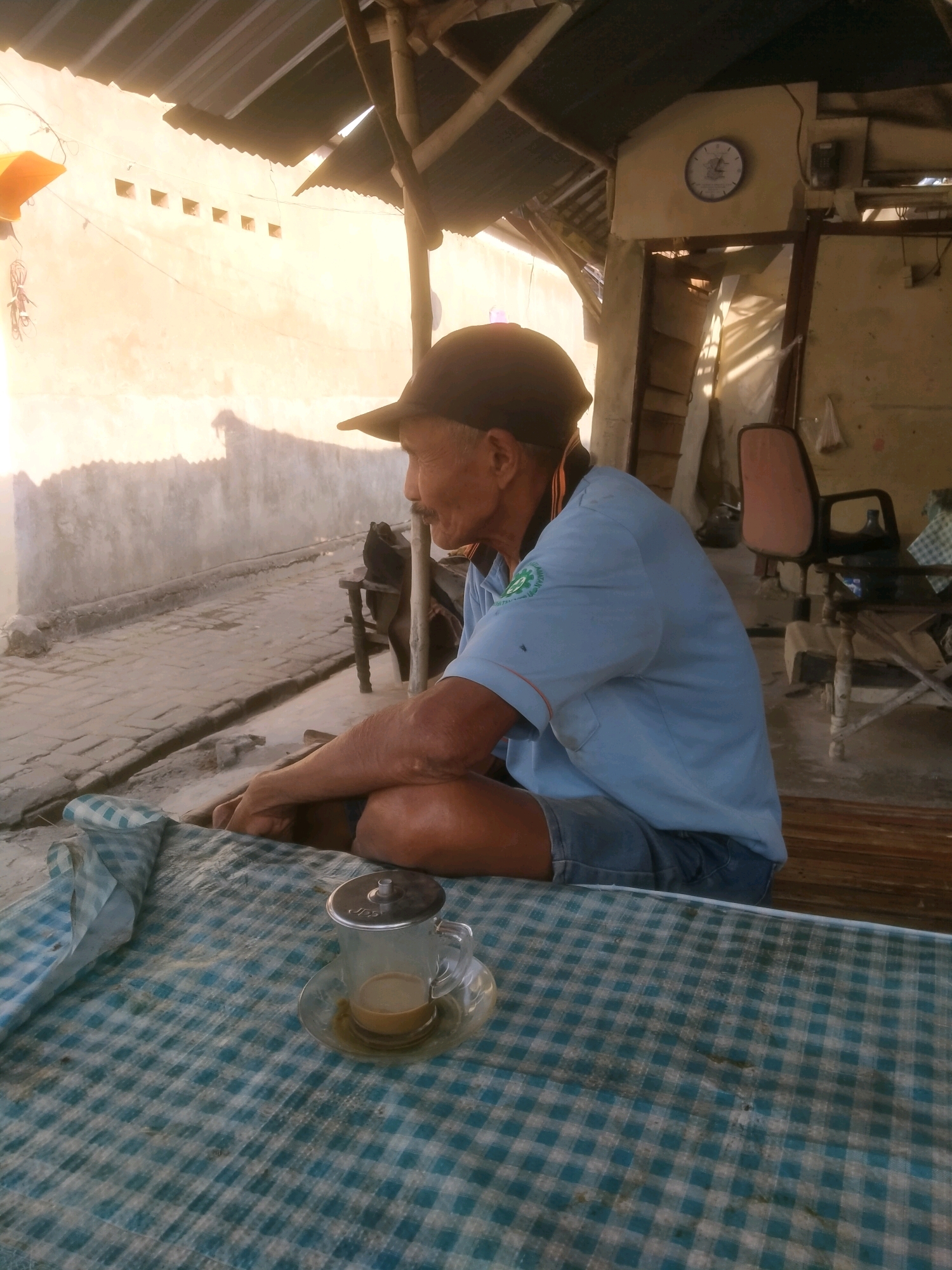 In Sumengko, Indonesia, an elderly man sits cross-legged on a wooden surface, facing away from the camera and looking into the distance. He wears a blue collared shirt with a green emblem on the left chest, denim shorts, and a black baseball cap. In front of him, a checkered blue and white cloth covers a table, on which rests a glass of light brown liquid with a metal lid and a saucer. The scene is set outdoors under a corrugated metal roof supported by bamboo poles. Sunlight illuminates a light-colored wall behind him, casting shadows. In the background, a clock hangs on the wall, and chairs and tables are visible, suggesting a small establishment. The ground is paved with bricks.