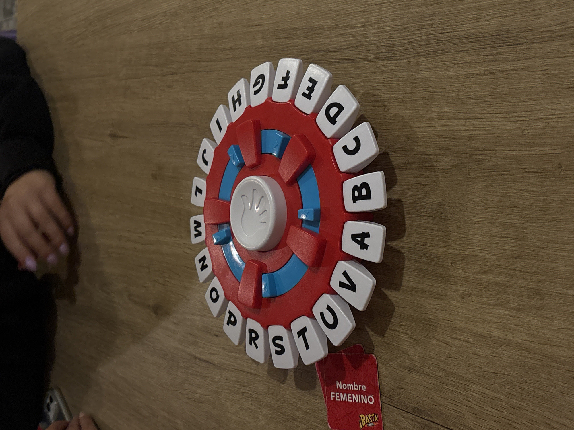 The image shows a red and blue circular game board with white letter tiles placed around its edge, spelling out the alphabet. A card labeled "Nombre FEMENINO" (Spanish for "Feminine Name") sits next to the game. Part of a person's arm and hand are visible on the left side of the image, and the scene is set on a light brown wooden surface. The location cannot be determined from the image.