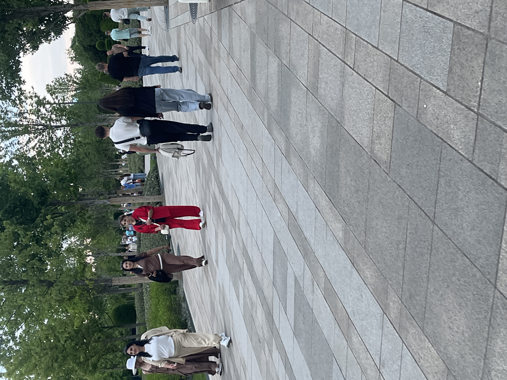 This rotated image captures a sunny, late afternoon or early evening scene in a public park or garden in Krasnodar, Russia. Numerous individuals are seen walking along a wide, paved pathway constructed from rectangular light grey and darker grey tiles.
In the foreground, a person in a white top and beige coat/dress is walking alongside another person in a brown dress, the latter wearing a white hat. Further back, a woman in a red pantsuit is seen, holding a white cup or bottle. Close to her, another woman in a long brown dress or abaya is walking with a black bag.
Behind these individuals, a group of people continues walking down the pathway. This group includes a person in a white polo shirt and black pants with a black sling bag, followed by someone with long dark hair in a black top and light blue jeans, and a person in a black t-shirt and blue jeans. Further into the background, other walkers are visible, including one in a light green polo shirt and another in a white top.
The pathway is bordered by lush green trees and manicured bushes, indicating a well-maintained outdoor environment. The sky appears clear, suggesting pleasant weather for a leisurely stroll. There is no visible text in the image. People appear relaxed and are engaged in casual walking, enjoying the outdoor setting.
