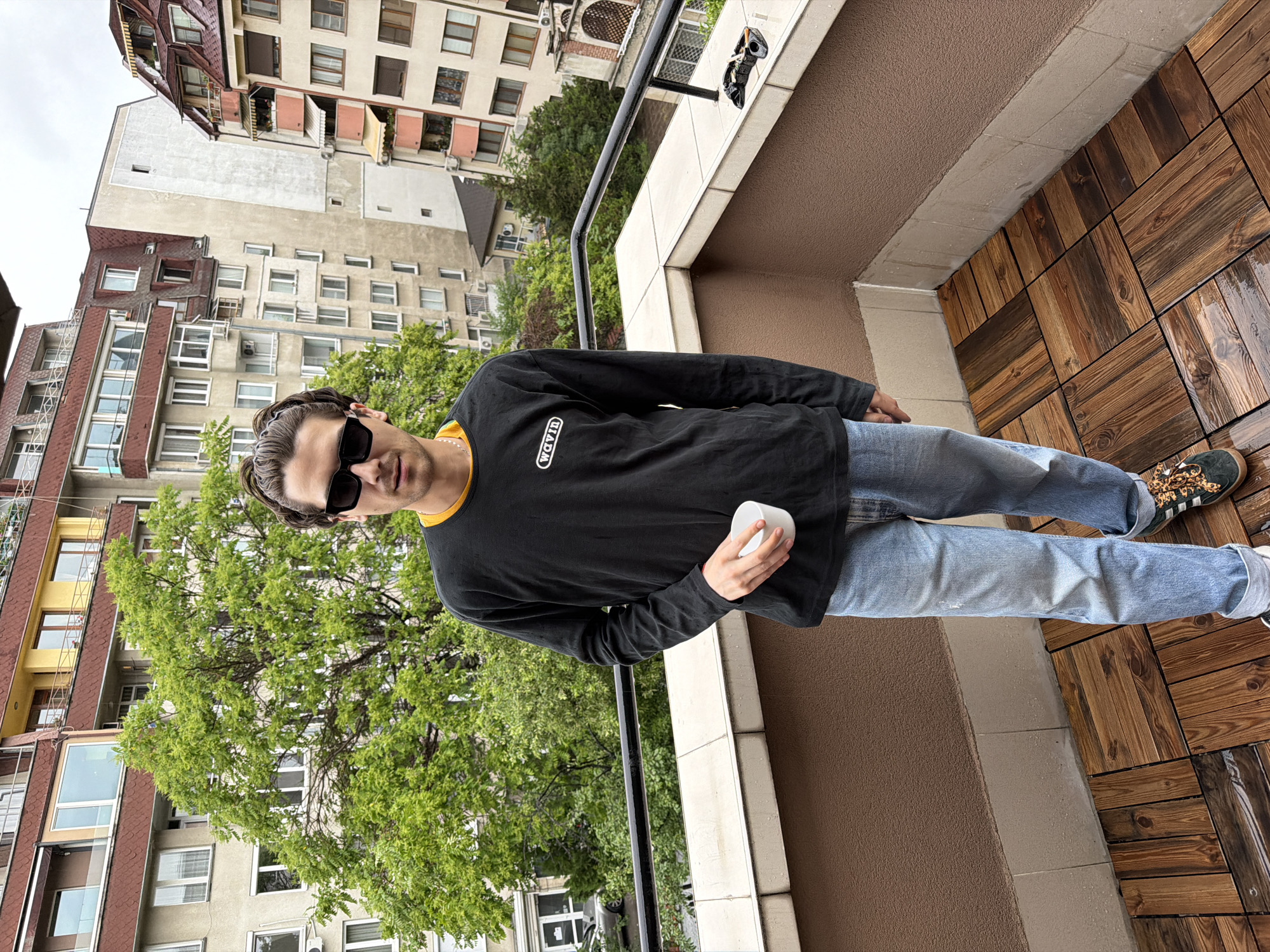 A young man stands on a balcony in Sofia, Bulgaria, looking casually towards the camera. He is wearing a black long-sleeved shirt with the word "WAVIN" in white text on the chest, a yellow undershirt collar, blue jeans, and leopard print sneakers with white stripes, possibly Adidas. Dark sunglasses obscure part of his face. In his left hand, he holds a small white cup or container.
The balcony features dark brown wooden floor tiles, which appear damp, suggesting recent rain. The balcony railing is black, and the immediate wall is a textured brown. In the background, an urban landscape unfolds with multiple residential buildings of varying heights and architectural styles, featuring beige and light-colored facades, some with reddish-brown roof sections and numerous windows. Lush green trees are visible between the buildings, adding a natural element to the city scene. The sky is overcast, indicating daytime with diffused light. The overall atmosphere is relaxed, capturing a moment on a balcony overlooking a bustling city.
