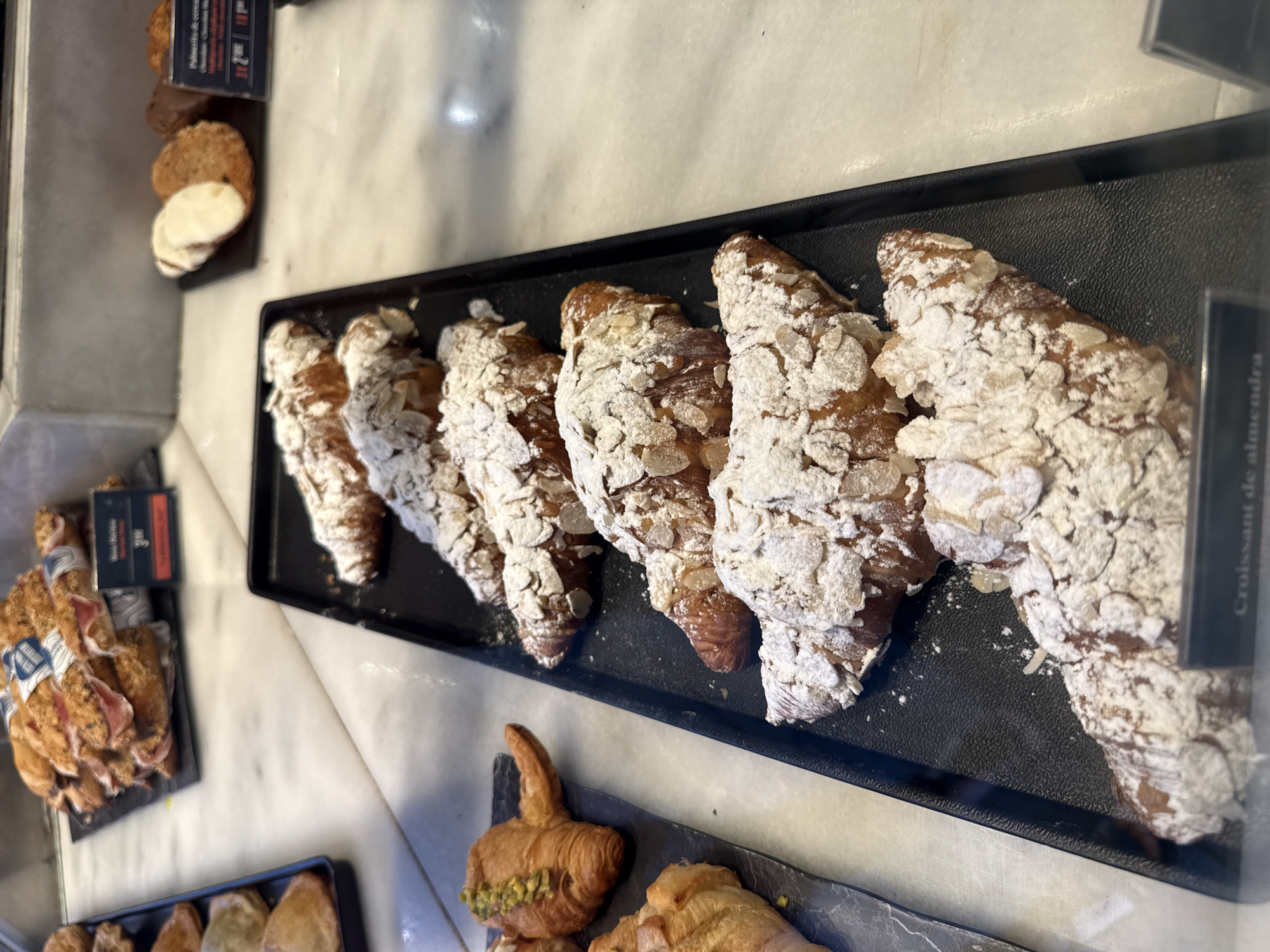 The image captures a close-up, slightly overhead view of a bakery or cafe display case, primarily featuring various baked goods. The central focus is a black rectangular tray filled with seven almond croissants ("Croissant de almendra"). These croissants are golden-brown, generously topped with powdered sugar and sliced almonds, and appear freshly baked. They are arranged in a row, slightly overlapping.
To the left of the almond croissants, on a separate small black tray, are several round, crumbly-looking cookies, some appearing to be split in half to reveal a light-colored interior. A price tag next to them reads "Polvorones de cerea" (Cereal shortbread cookies) and "3 / 2€," indicating three units for two Euros.
Further to the left, another black tray holds pastries that resemble bundles of thin, flaky dough wrapped around a filling, possibly savory like ham, as a partially visible label next to them reads "MASA HOJALDRE" (Puff pastry dough) and "JAMON" (Ham), with a price of "3€."
In the foreground, another black or dark gray tray displays more pastries, including one that appears to be a croissant with a green filling, possibly pistachio, and other unidentifiable baked items.
The setting features light-colored, possibly marble, countertops or surfaces. The overall scene suggests a well-lit, commercial environment dedicated to selling pastries and baked goods. The visible Spanish text ("Croissant de almendra," "Polvorones de cerea," "Masa hojaldre," "Jamon") indicates a Spanish-speaking location.