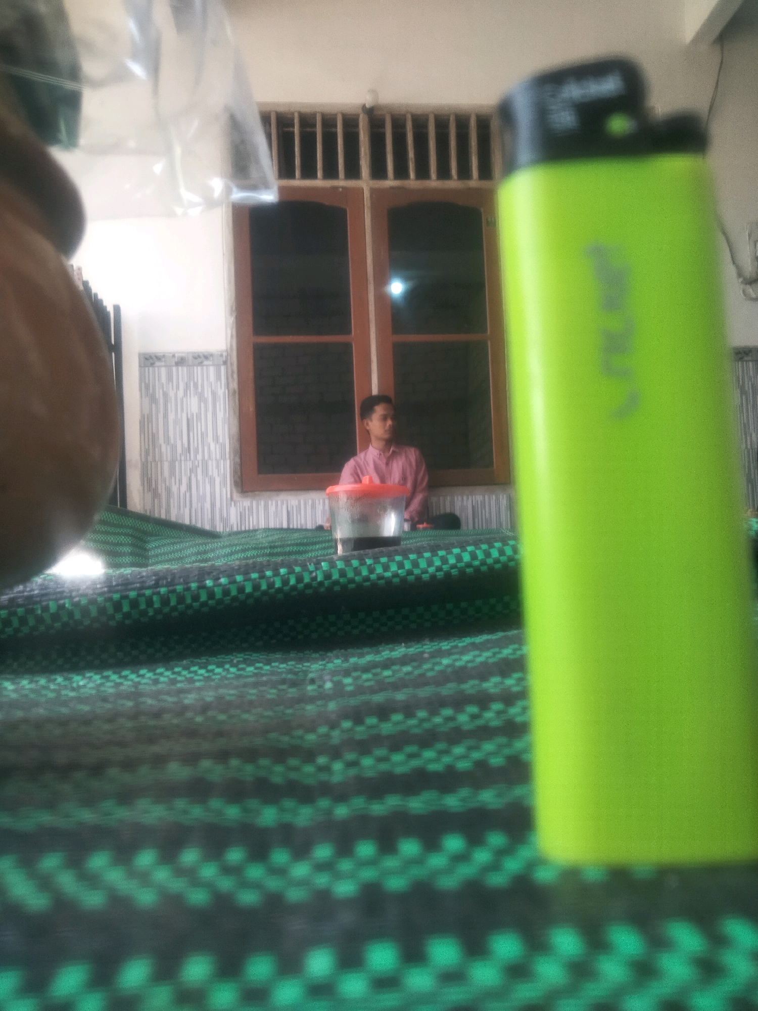In a residential setting in Soko, Indonesia, a close-up shot focuses on a bright green lighter in the foreground, obscuring a portion of the background. The lighter has black accents on its top. Behind the lighter, a woven mat with a checkered pattern of green and black covers the surface. Further back, a young man wearing a pink collared shirt is seated. He is looking towards the left of the frame. Next to him is a clear plastic cup with a red lid. The background features a window with dark panes and wooden frames, flanked by vertical bars above. The wall behind the window has a textured tile pattern. A plastic bag is visible hanging in the upper left of the image, partially obscuring the ceiling and wall.