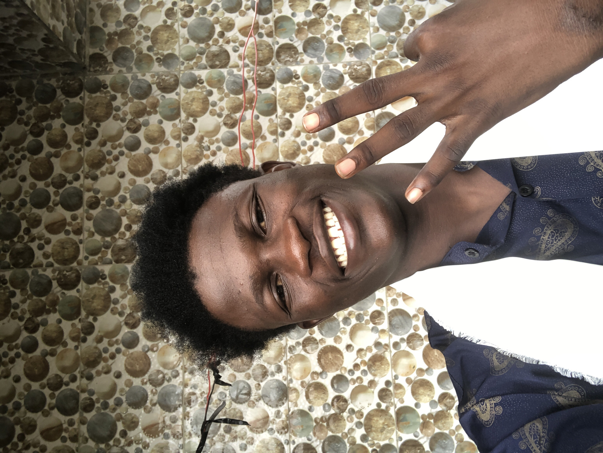 A close-up, waist-up, self-portrait shows a young Black male with dark skin, dark curly hair, and a wide smile. He is wearing a dark blue shirt with a gold paisley pattern and a white cloth draped around his neck. His right hand is raised, displaying a peace sign with his index and middle fingers. The background consists of a tiled wall with a circular mosaic pattern in shades of brown, beige, and grey. Two red wires are visible strung horizontally across the upper portion of the wall, and black ribbon-like material hangs on the left side of the frame. The lighting is bright, casting a slight shadow on the left side of the man's face. The overall scene appears to be outdoors, possibly in a residential or public area in Ado, Nigeria, given the typical use of such tiling in the region.