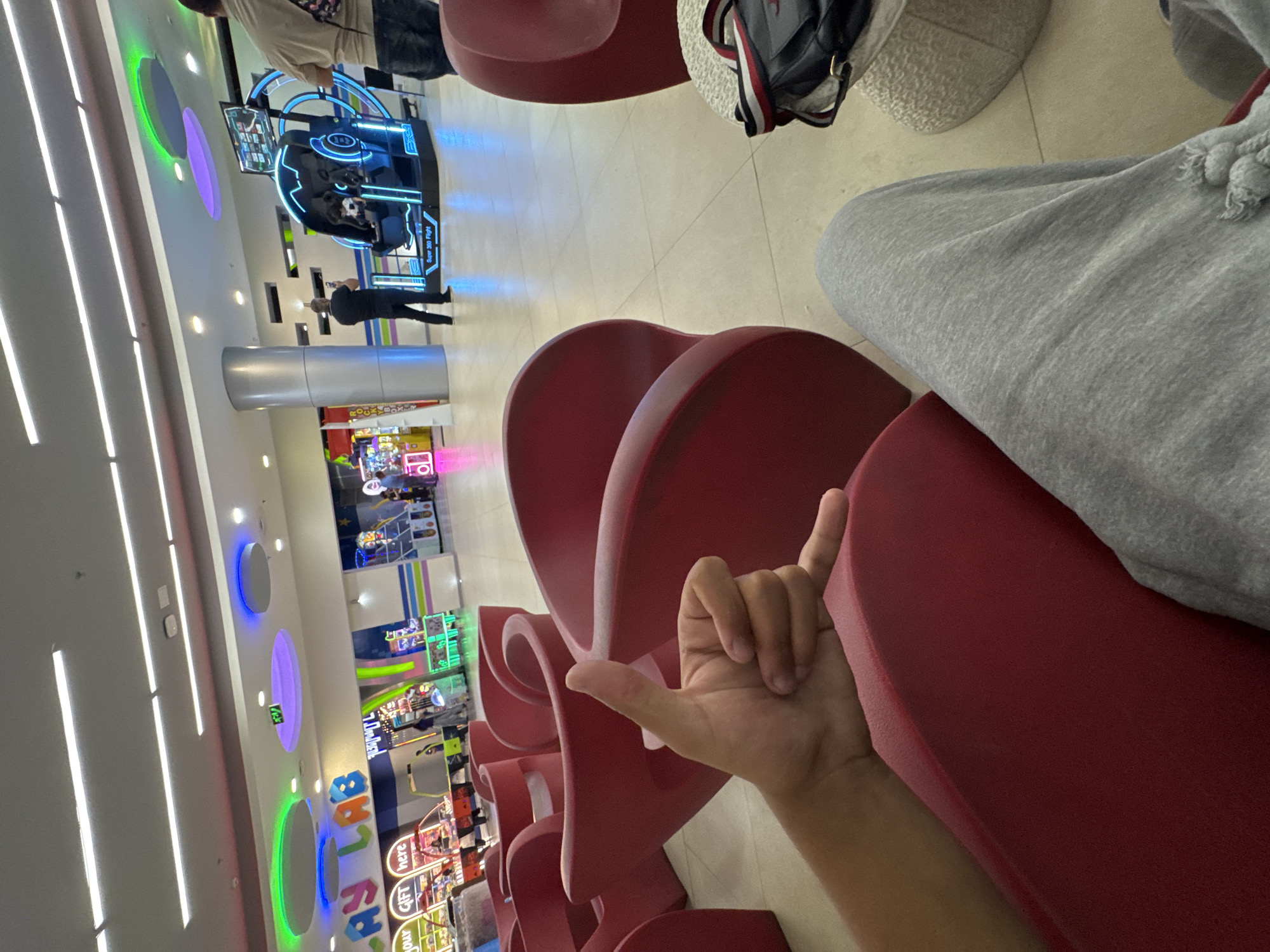 This media file depicts an indoor scene within a modern entertainment or arcade center, likely in Yerevan, Armenia. The perspective is from a seated position, looking towards the activity in the room.
In the foreground, a person's arm and hand are visible, making a "shaka" gesture, resting against a red, sculpted chair. Adjacent to this, part of a person's leg, clad in grey sweatpants with a visible drawstring, is also seen, suggesting another individual is seated nearby. A black backpack with red and white stripes is placed on a light-colored, textured stool or small table, close to the seating area.
The background unfolds into a brightly illuminated entertainment hall. The ceiling features numerous long, white linear lights and several circular light fixtures emitting green, purple, and blue glows, contributing to a vibrant atmosphere. Along the walls, various arcade games and attractions are present. A prominent feature is a "Super 360 Flight" simulator, identified by its blue neon lighting and interactive setup. A person is visible interacting with the simulator, while another individual in dark clothing stands nearby. Further back, another person in a light-colored shirt is seen near other games. On the left side, closer to the foreground, various arcade machines are visible, including one with text "Your GIFT here." Partial letters of a large, colorful sign, possibly spelling "PLAY" or "TOYS," are also discernible.
The scene captures an active indoor environment, with people engaged in or waiting for various entertainment activities. The "shaka" sign in the foreground suggests a relaxed and friendly mood. The lighting and array of games indicate a typical family entertainment center or arcade.