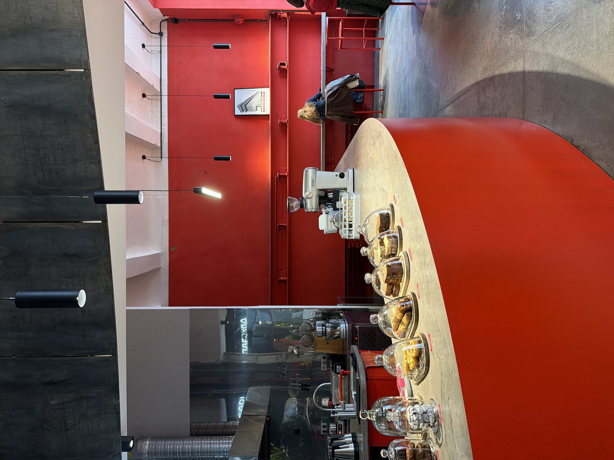 An interior shot captures a modern cafe or bar with a striking industrial aesthetic, dominated by vibrant red and cool grey tones. The view is slightly angled, looking towards a long, curved counter that forms a prominent feature.
One person with blonde hair, wearing a dark jacket, is seated at a red table or counter in the mid-ground, facing away from the viewer. Another person is partially visible in the upper right corner, wearing a red top.
The cafe features a bright red wall with visible industrial beams and piping. Several black cylindrical pendant lights hang from the ceiling, with at least one emitting a bright white light. On the red wall, a framed picture displays a building and the Cyrillic word "МОСКВА" (Moscow), confirming the location.
The grey counter, possibly concrete or a similar material, showcases an array of pastries and desserts displayed under clear glass cloches. A silver-toned coffee machine and grinder are also visible on the counter. The floor is a textured grey, resembling concrete, with subtle lines and shadows. Exposed ductwork and other industrial elements are seen in the lower left section, near a dark, reflective surface that shows a blurred reflection of "ПАРХОД" (possibly "ПАРОХОД" meaning "steamboat" or "steamship"), likely from a sign within the establishment. The overall atmosphere appears calm and contemporary.