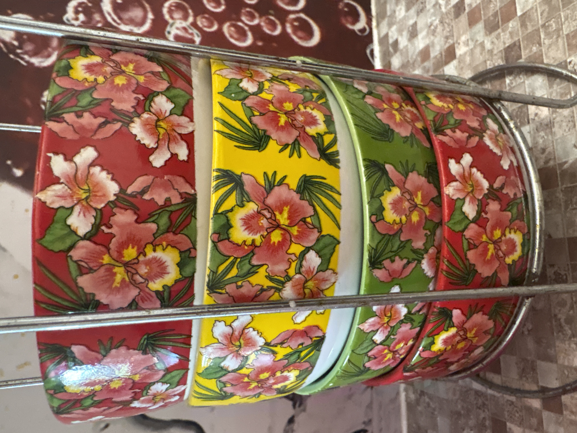 Four floral-patterned ceramic bowls are stacked in a metal rack. The bowls have a red, yellow, green, and red exterior, respectively, each decorated with pink and yellow orchids and green leaves. The rack is metallic and appears to be wire-formed. The bowls are on a speckled countertop. To the right of the bowls, a dark brown surface with lighter brown circular patterns is visible.