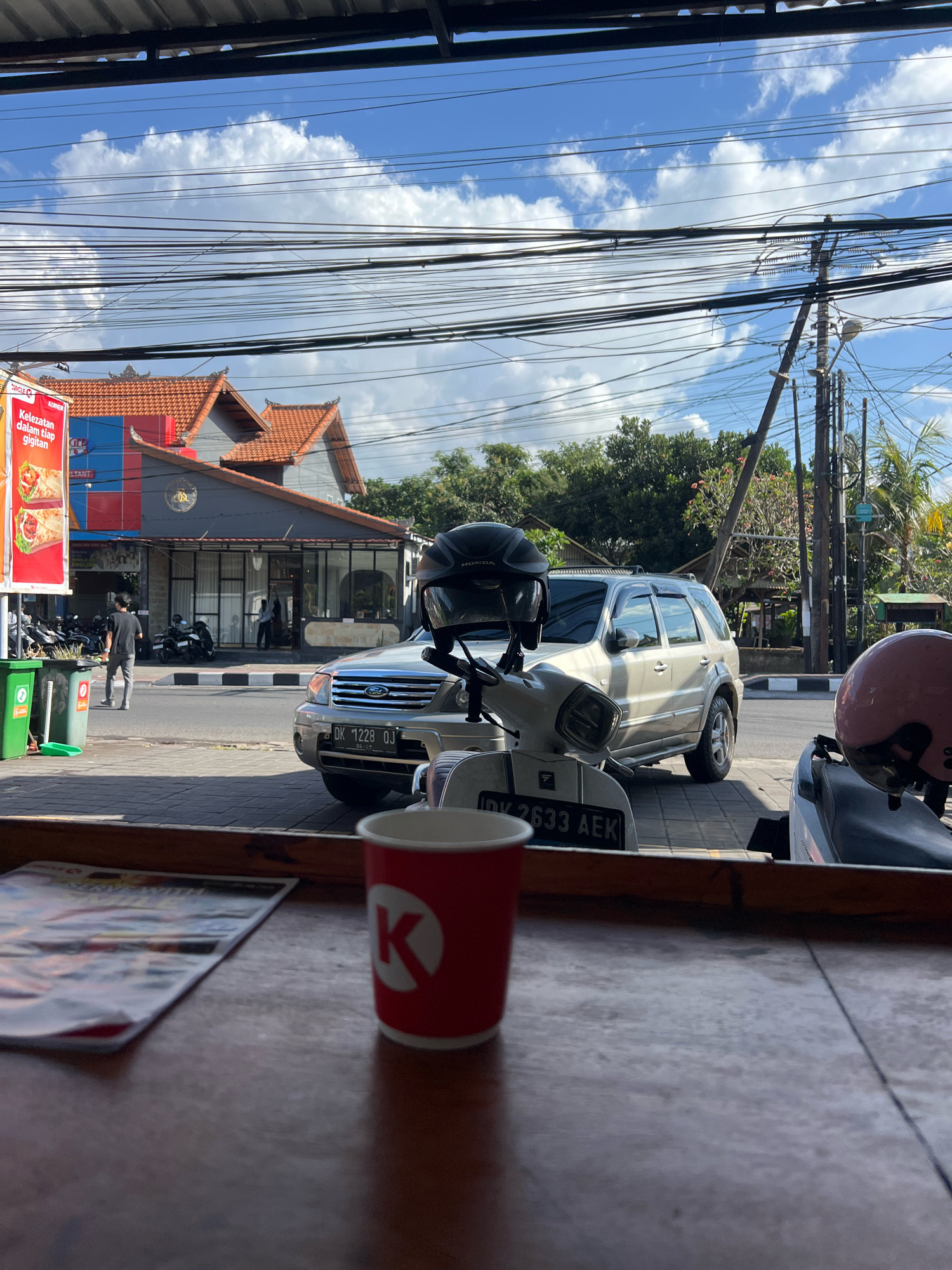 In Denpasar, Indonesia, a street-level view captures a scene outside a building with a red-tiled roof. A white SUV, license plate DK 1228 OJ, is parked on the asphalt. In front of it, a white scooter is parked, adorned with a black helmet. To the right, another scooter, partially visible, features a pink helmet. A red cup with a white circular logo, likely for coffee, sits on a wooden surface in the foreground. A magazine lies beside the cup, also on the wooden surface. Across the street, a man in a black t-shirt walks past green trash cans. The sky above is blue, dotted with white clouds, and crisscrossed by a dense network of electrical wires.
