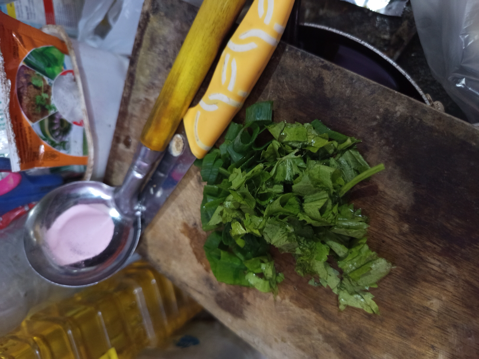 The image depicts a cutting board with chopped green vegetables, likely scallions and cilantro, situated in what appears to be a kitchen. A metal ladle with a pinkish substance inside is positioned above the cutting board. To the right of the cutting board are two knives with yellow handles. In the background, a bottle of cooking oil and packages of food products are visible. The scene is likely a domestic setting in Tebel, Indonesia, given the context provided. There are no people visible in the image, and no specific actions or interactions are being captured.