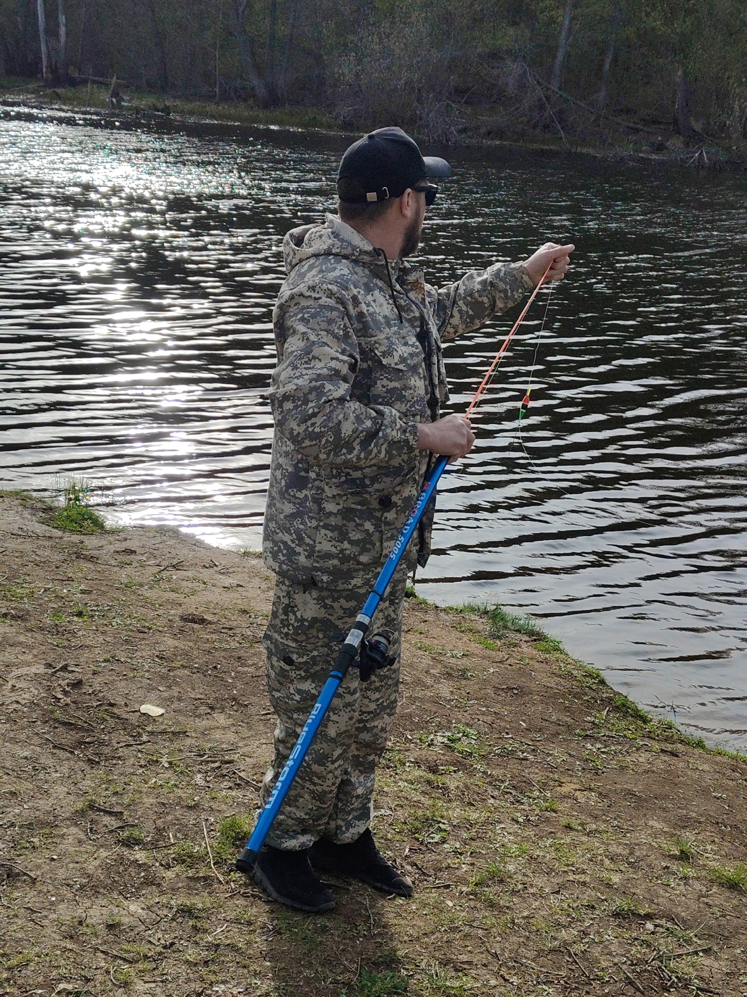 A person is fishing near a body of water, possibly a river or lake. The individual is wearing a camouflage jacket and pants, along with a black baseball cap. They are holding a blue fishing rod and casting a line into the water. The water reflects sunlight, creating a shimmering effect. The person is standing on a grassy bank, with trees visible in the background. The location is unspecified.