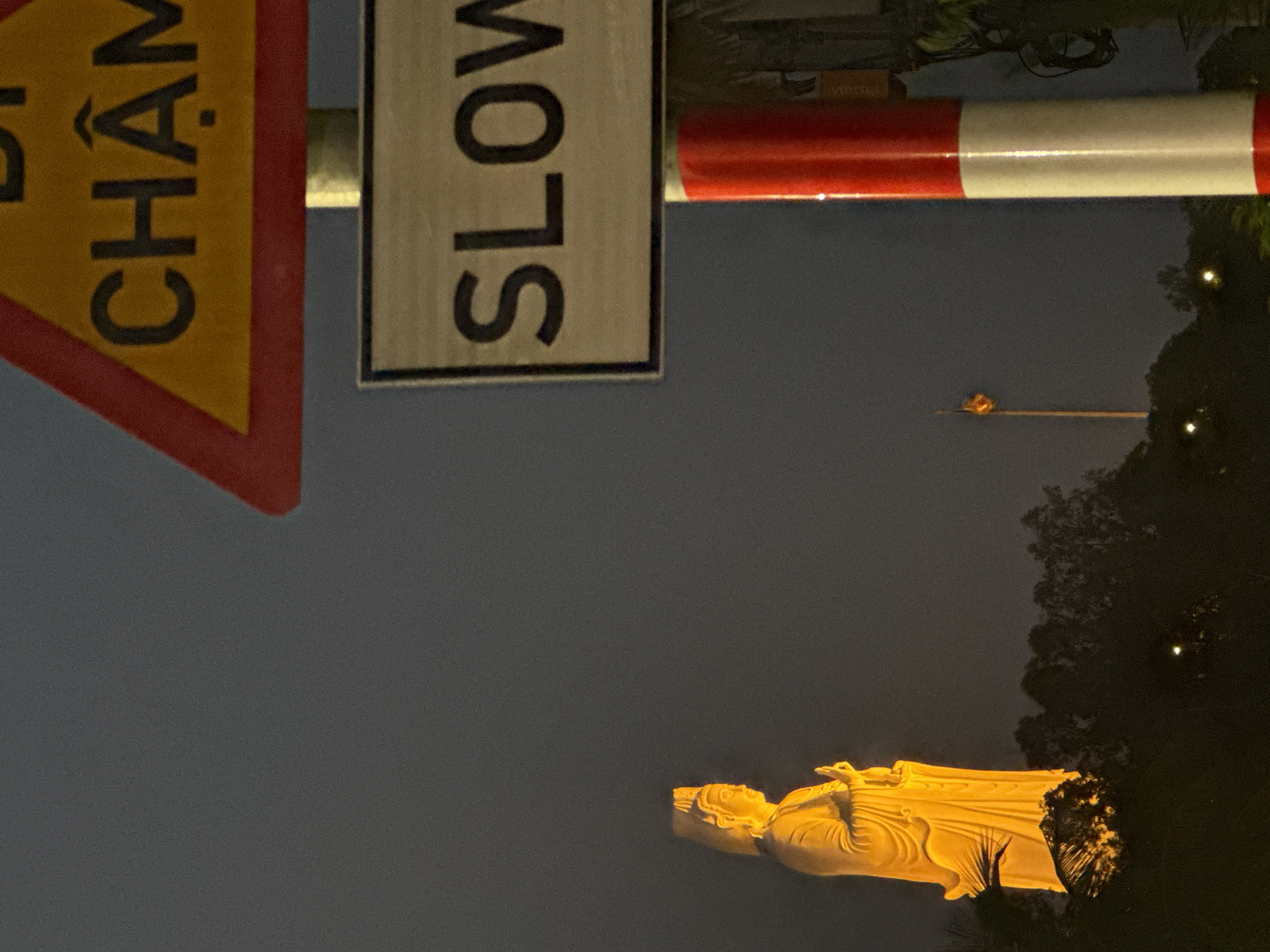 The image shows a nighttime view of the reclining Buddha statue at the Linh Ứng Temple in Đà Nẵng, Vietnam. In the foreground are traffic signs, one triangular with Vietnamese script and the other a rectangular "SLOW" sign. A red and white traffic barrier is visible nearby. The illuminated Buddha statue is in the lower part of the image, with trees and the night sky in the background. A tower or structure with lights can be seen in the distance.