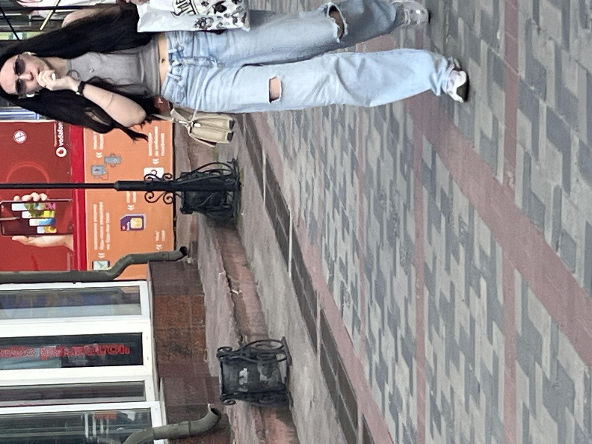 The image shows a young woman walking on a sidewalk in Dnipro, Ukraine. She is wearing ripped light-wash jeans, a grey tank top, white sneakers, and sunglasses. She is holding a white shopping bag in her right hand and a beige purse on her left shoulder. She appears to be eating something from her right hand, which is near her mouth. Behind her, there is a red billboard with some text and images, and a street lamp. To her left, there is a decorative metal trash can. The sidewalk is paved with a pattern of grey and red interlocking stones. The overall scene suggests an ordinary day in an urban environment.