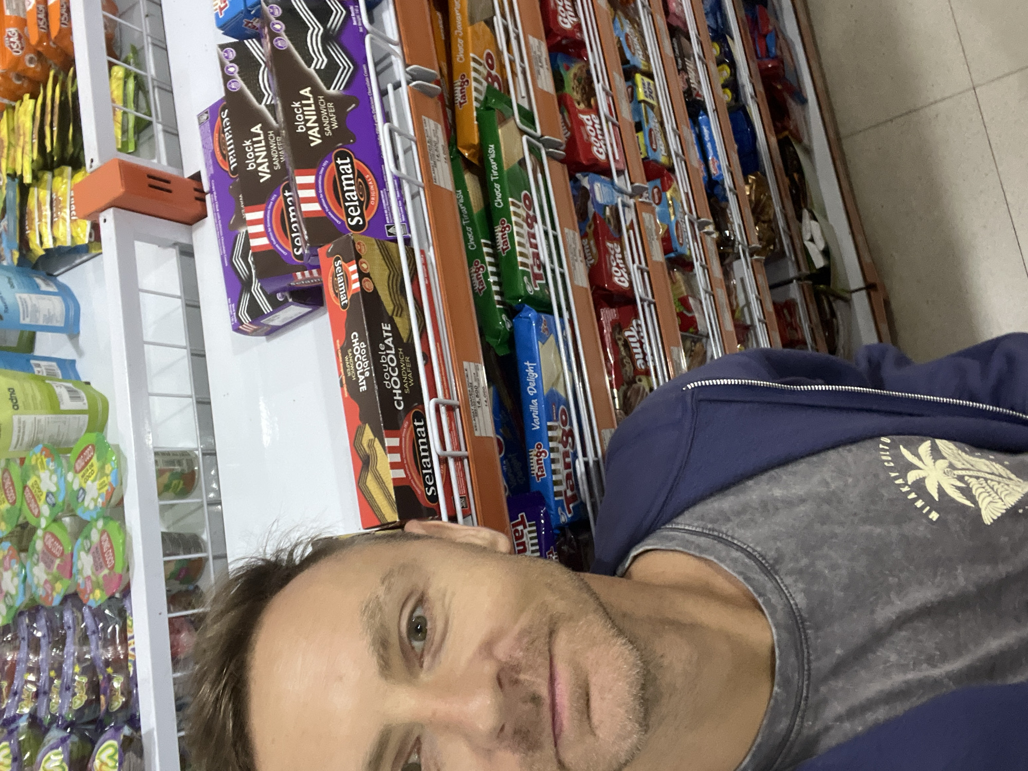 The image shows a selfie taken by a fair-skinned man with short, brown hair, wearing a gray t-shirt with a graphic design and a navy blue zippered jacket. He is standing in front of a shelf in what appears to be a convenience store or supermarket. The shelves are filled with various packaged snacks and candies, including brands like "Selamat," "Tango," and "Good Time." The "Selamat" brand is prominently displayed, featuring items such as "black Vanilla sandwich wafer" and "double Chocolate sandwich wafer". Other items include candies in green wrappers labeled "WONG COCO", "Usagi", and "Tango Choco Tiramisu". The environment suggests a retail setting, possibly in the city of Sukawati, Indonesia, given the product brands.