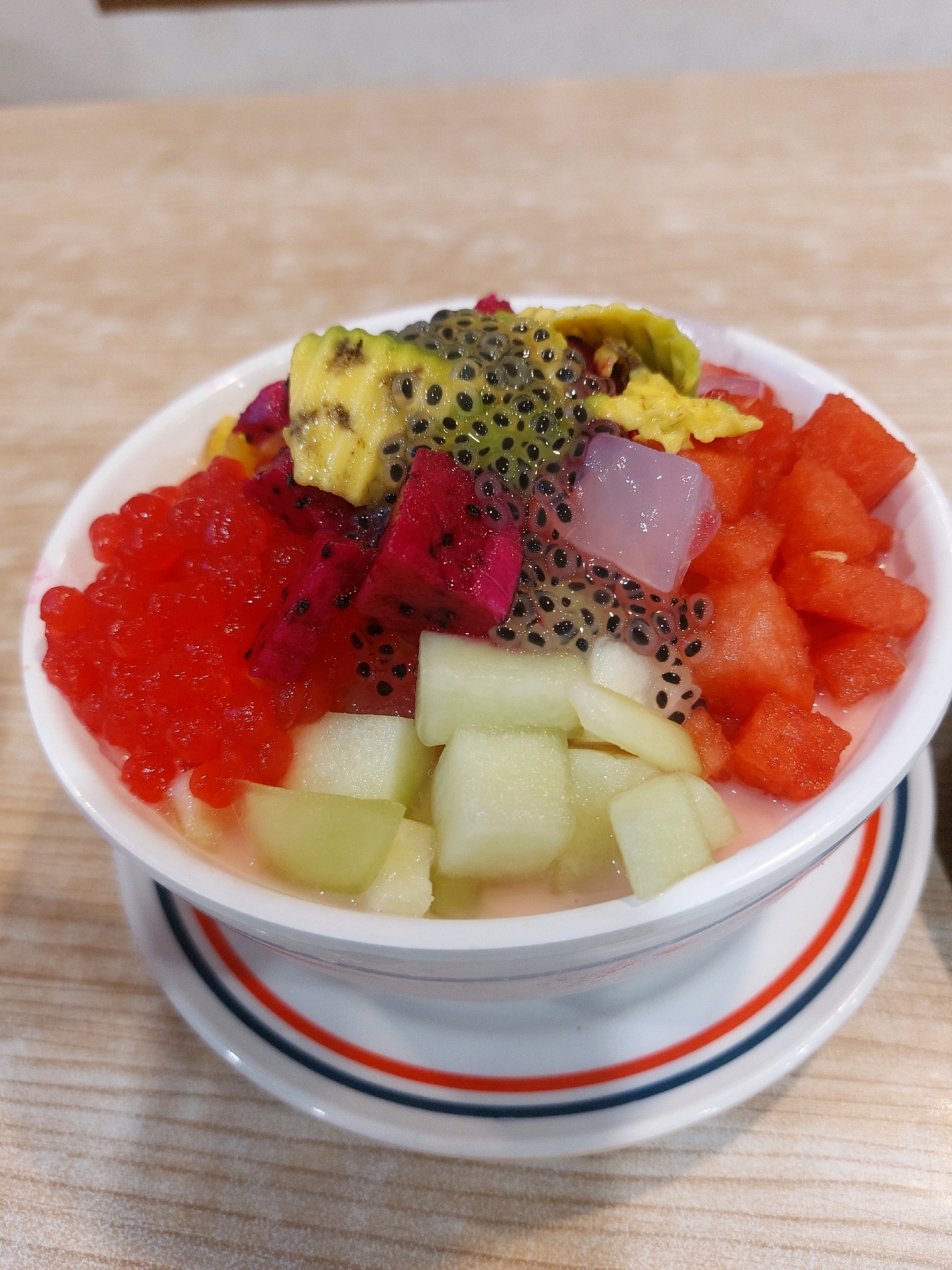 A white bowl filled with a colorful fruit dessert sits on a saucer with red and blue stripes. The dessert contains various fruits, including diced watermelon, dragon fruit, cantaloupe, avocado, and jelly. Some of the fruit is topped with basil seeds and a clear liquid. The bowl and saucer are placed on a light-colored wooden table. The image appears to be taken at a restaurant or food stall in Tarakan, Indonesia, given the context provided by the user.