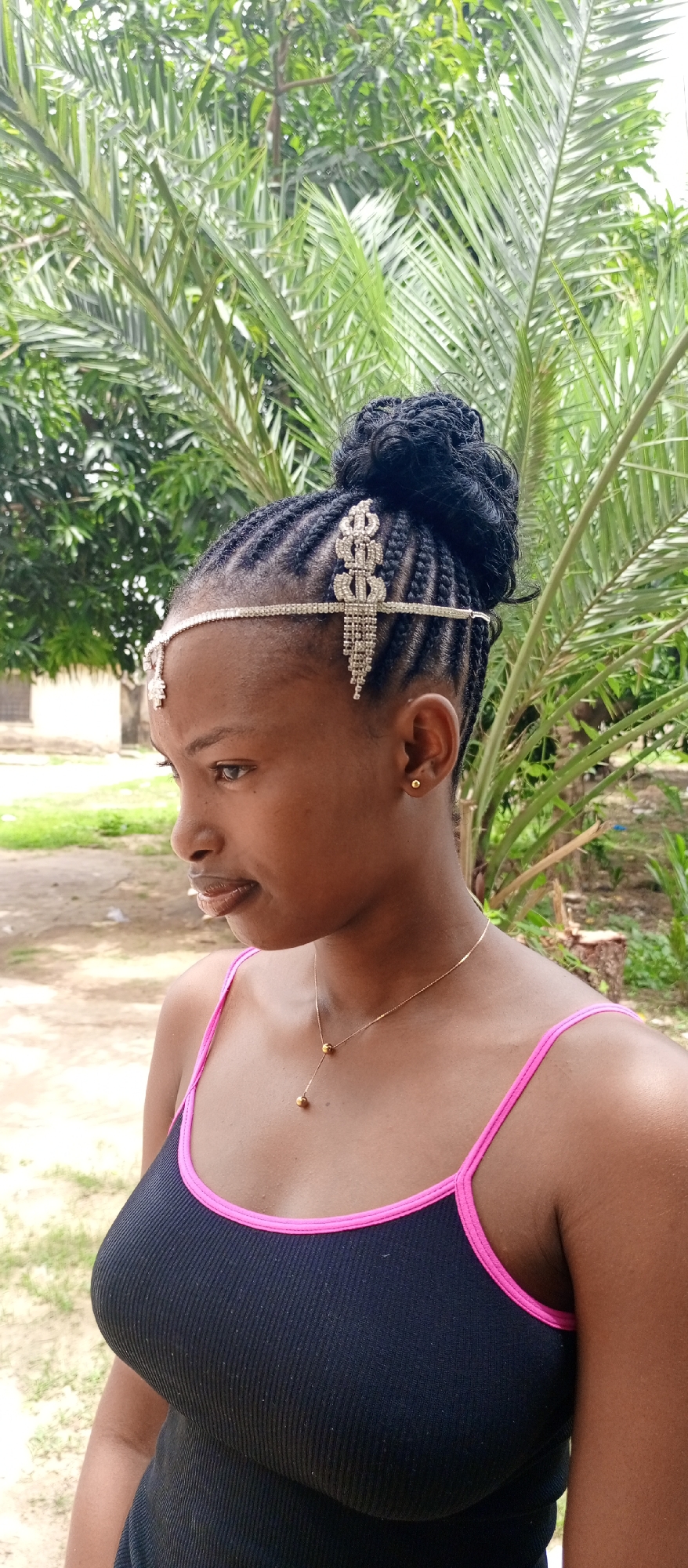 A medium close-up portrait shows a young woman, likely in Keffi, Nigeria, captured outdoors during daylight hours. She is depicted in profile, looking towards the left side of the frame with a neutral, possibly contemplative, expression. Her hair is intricately styled in cornrows that lead into a prominent bun or updo at the crown of her head. A decorative, sparkling silver-toned headpiece adorns her forehead and hair, featuring an ornate design with dangling rhinestone chains over her right temple.
She wears a simple gold stud earring in her visible right ear. A delicate gold chain necklace, featuring two small gold spherical beads, hangs around her neck. Her attire consists of a black ribbed tank top with distinct bright pink trim along the neckline and shoulder straps. The background is characterized by abundant green foliage, predominantly large, frond-like leaves indicative of palm trees or similar tropical vegetation. The natural lighting suggests a clear day. No visible text is present in the image.
